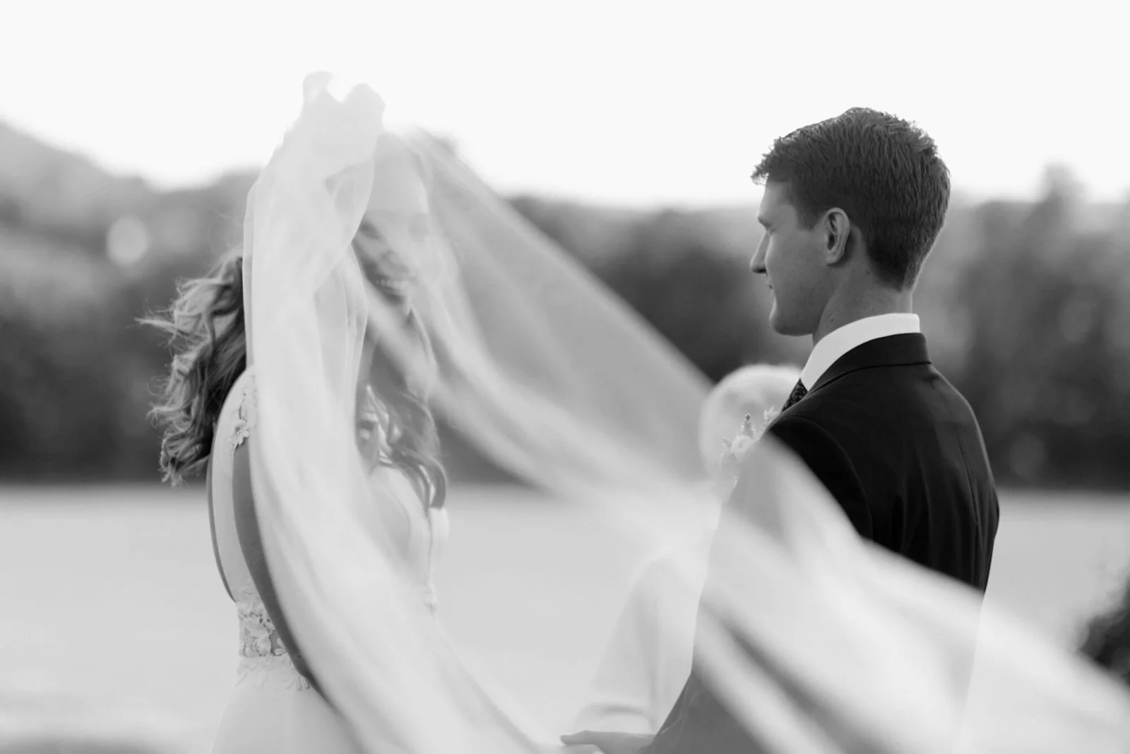 Black and white photo of a bride and groom on their wedding day, outdoors, with the bride's veil flowing between them.