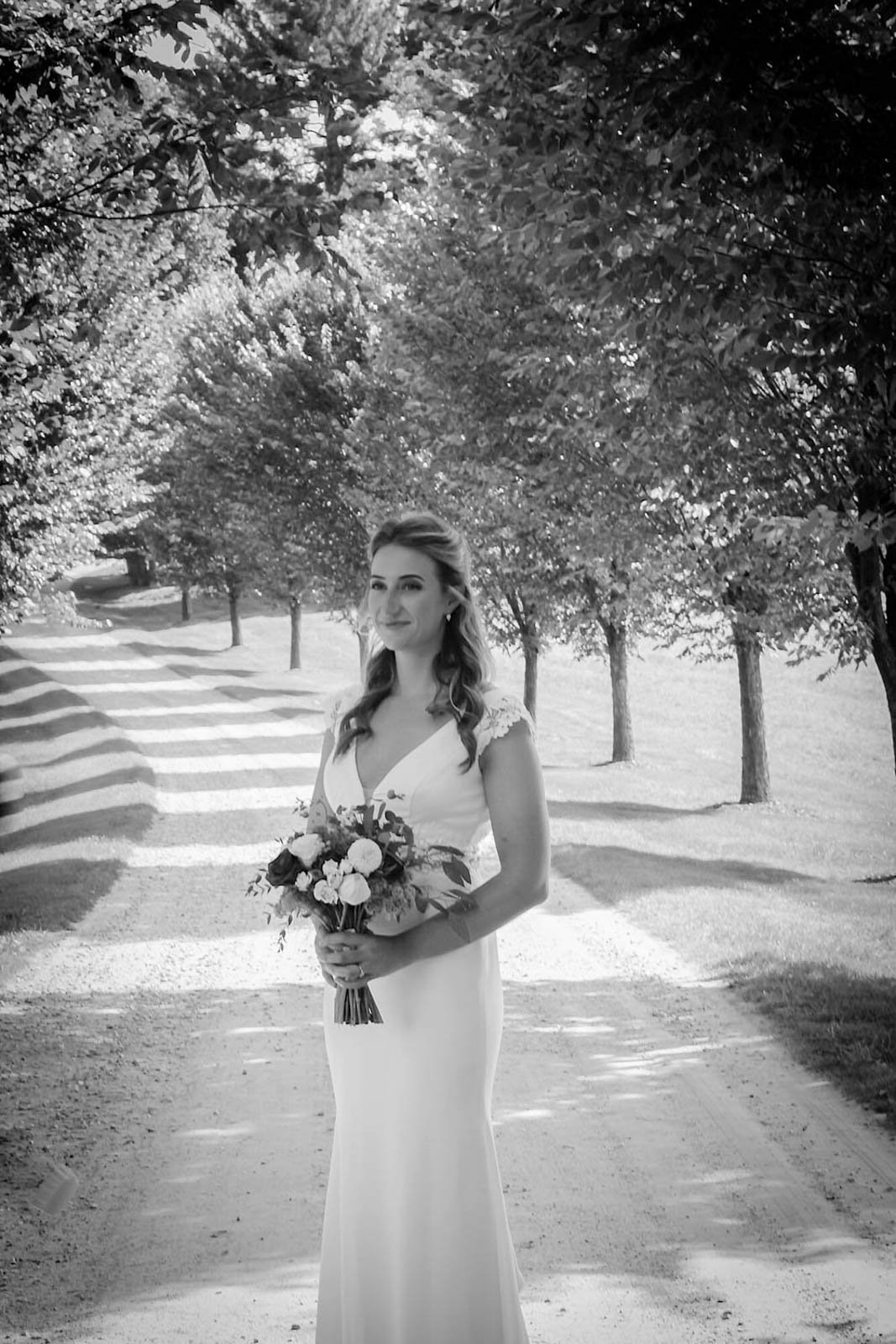 Black and white photo of a woman in a wedding dress holding a bouquet, standing on a dirt path lined with trees.