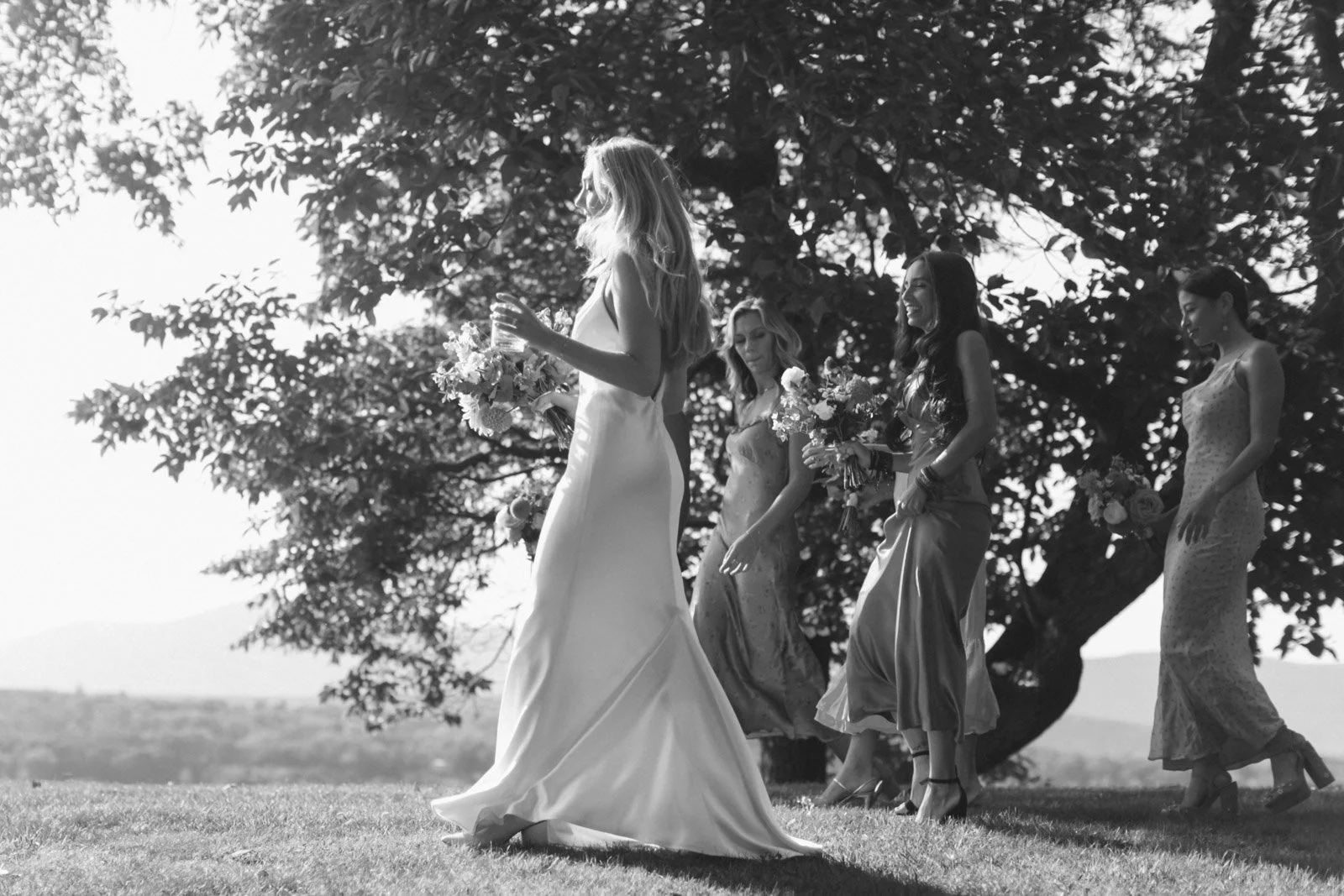 Black and white photo of a bride in a wedding gown walking on a grassy field with four women in dresses, holding bouquets of flowers, near a large tree with branches and leaves, and a landscape with hills in the background.