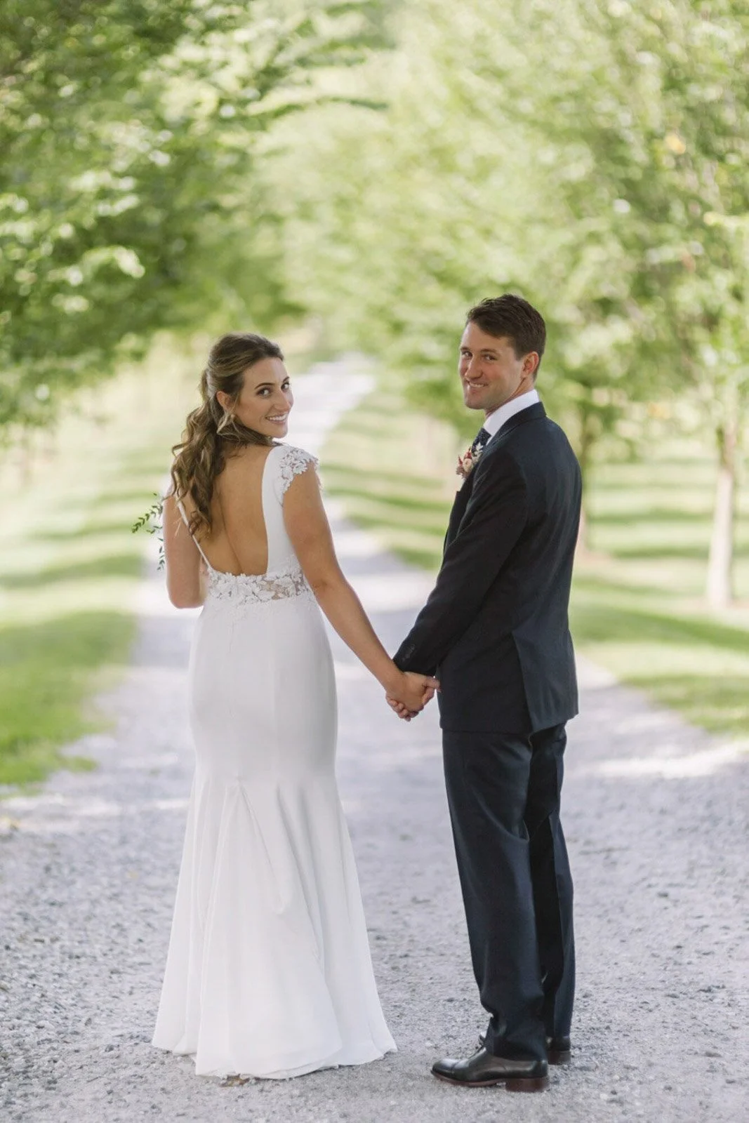 A bride and groom in wedding attire holding hands outdoors on a gravel path, with trees and greenery in the background, smiling at the camera at Court Hill in The Berkshires.