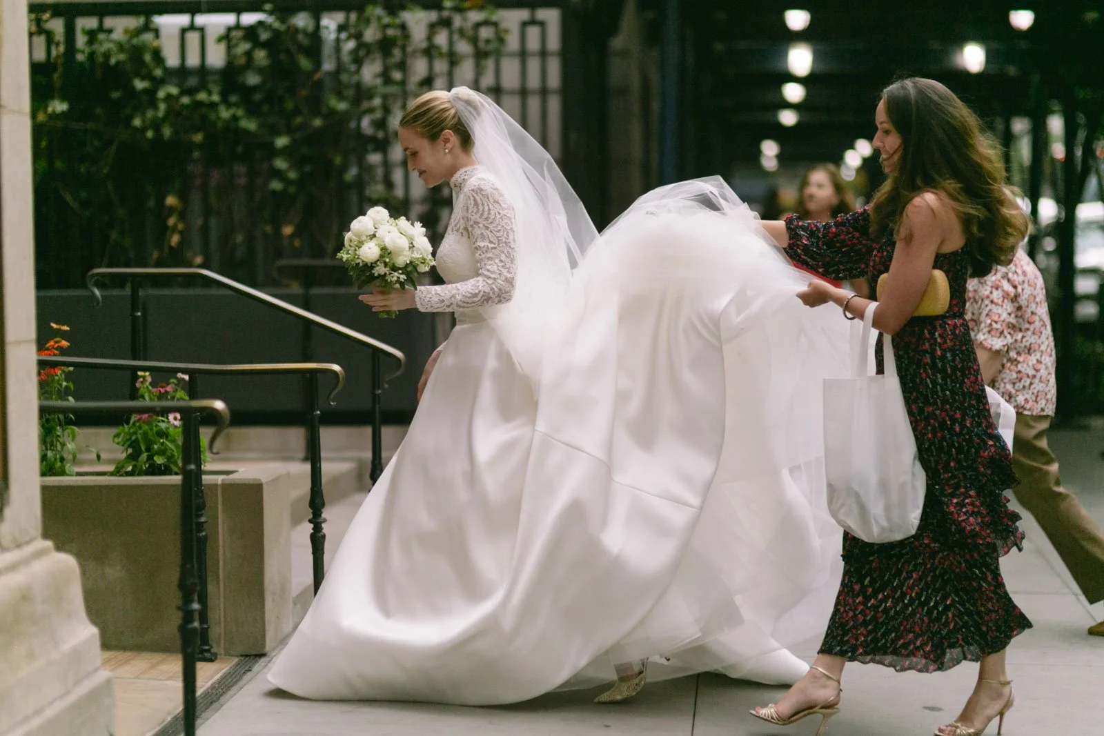 A bride in a white wedding gown and veil is being assisted by a woman in a black dress with red floral pattern as she walks down a city sidewalk. The bride holds a bouquet of white flowers.