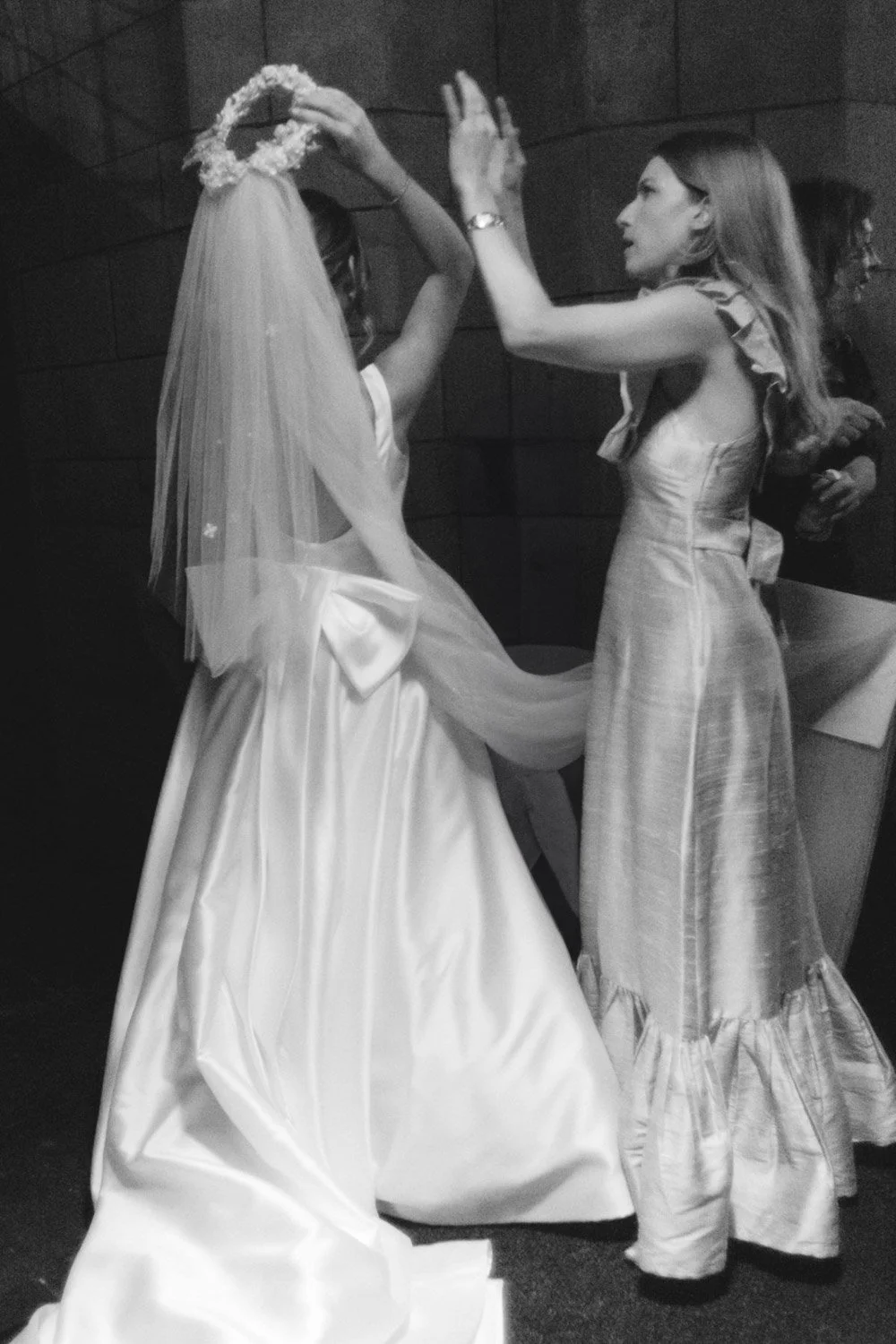 Two women in formal dresses, one adjusting a bridal veil on the other, in a dimly lit room or hallway.