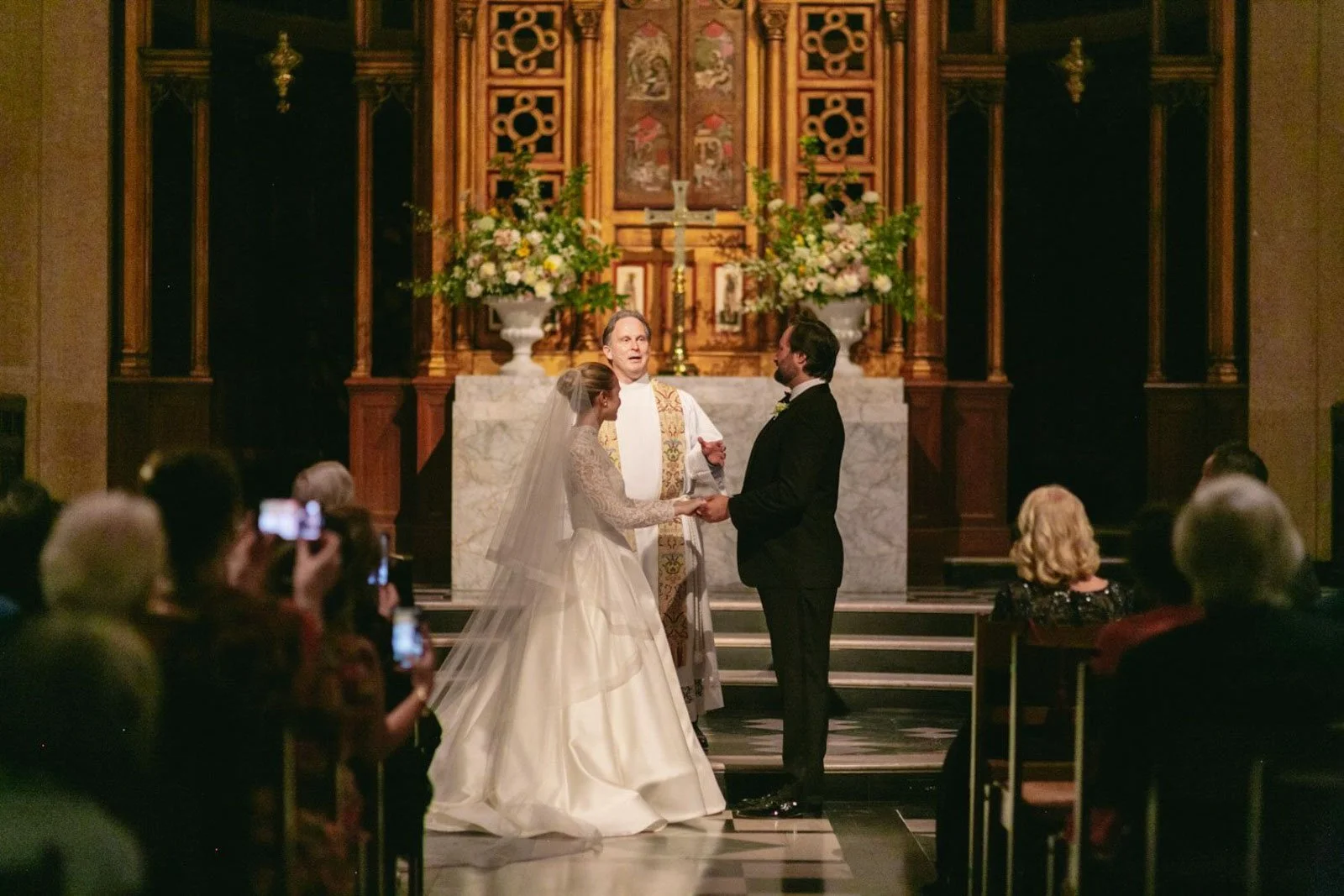 A wedding ceremony inside a church with a bride and groom standing before a priest, exchanging vows, with guests seated and capturing the moment.
