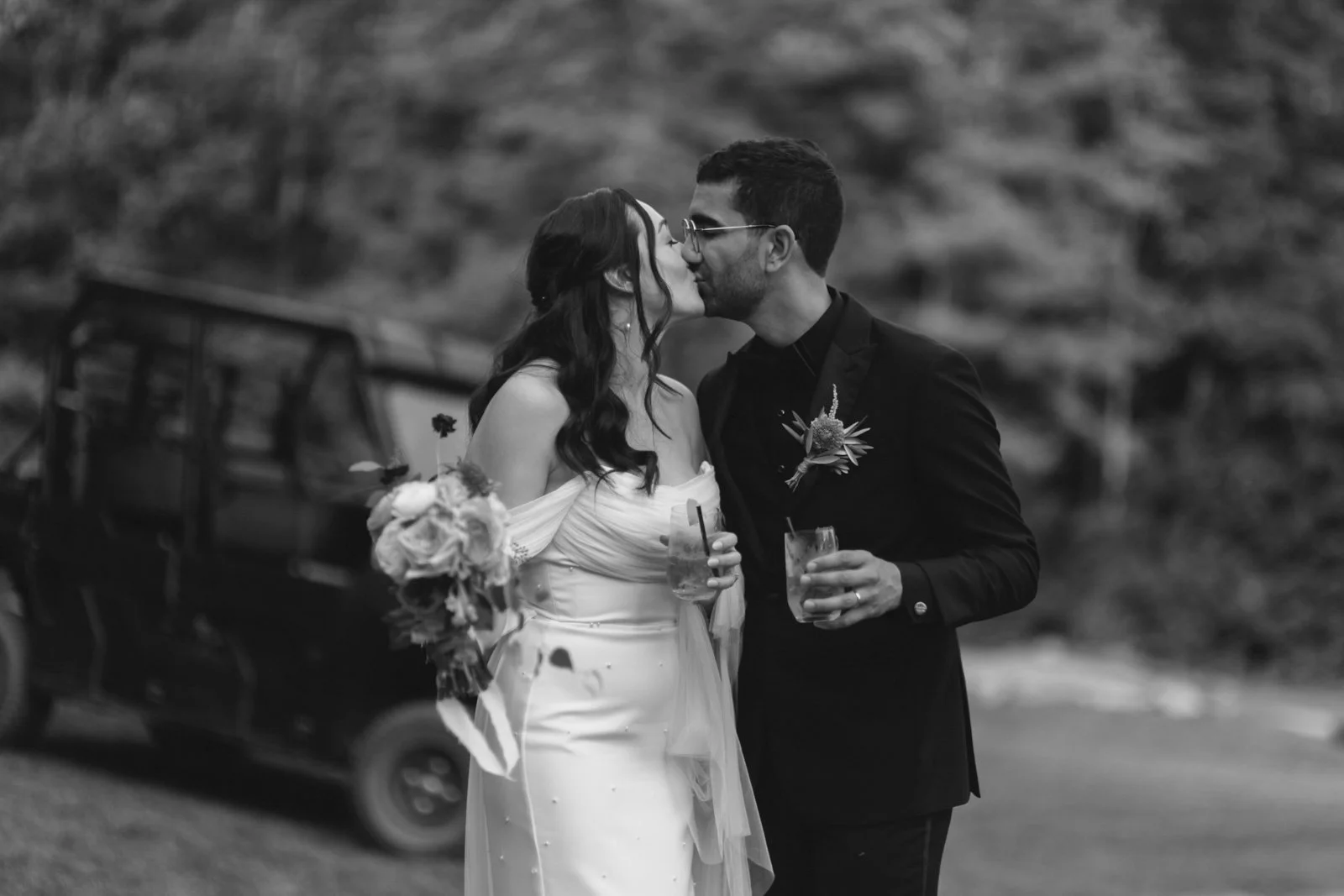 Black and white photo of a bride and groom kissing, holding drinks, with a car and hilly landscape in the background.