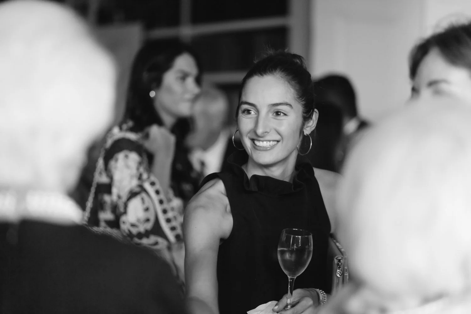 A woman smiling at a social gathering, holding a glass of wine, with other people around her at a wedding rehearsal dinner at The River Club in NYC.