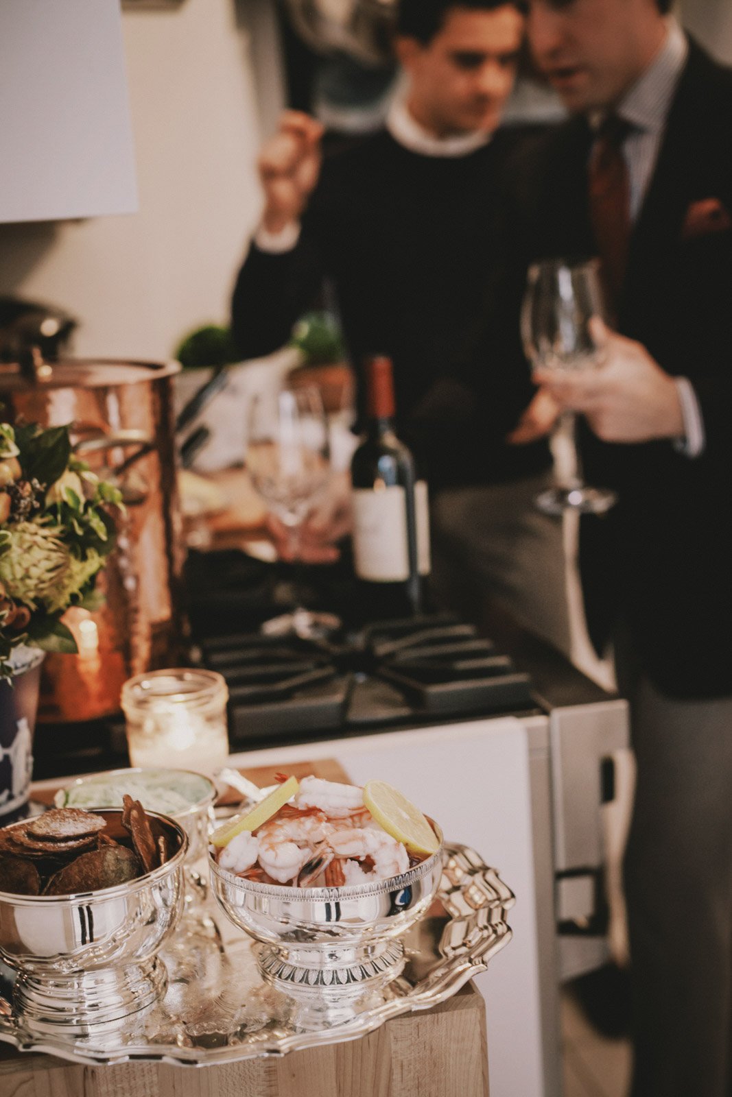 Dish with shrimp and lemon wedges on a silver tray in front of two men holding glasses, at a social gathering or party.
