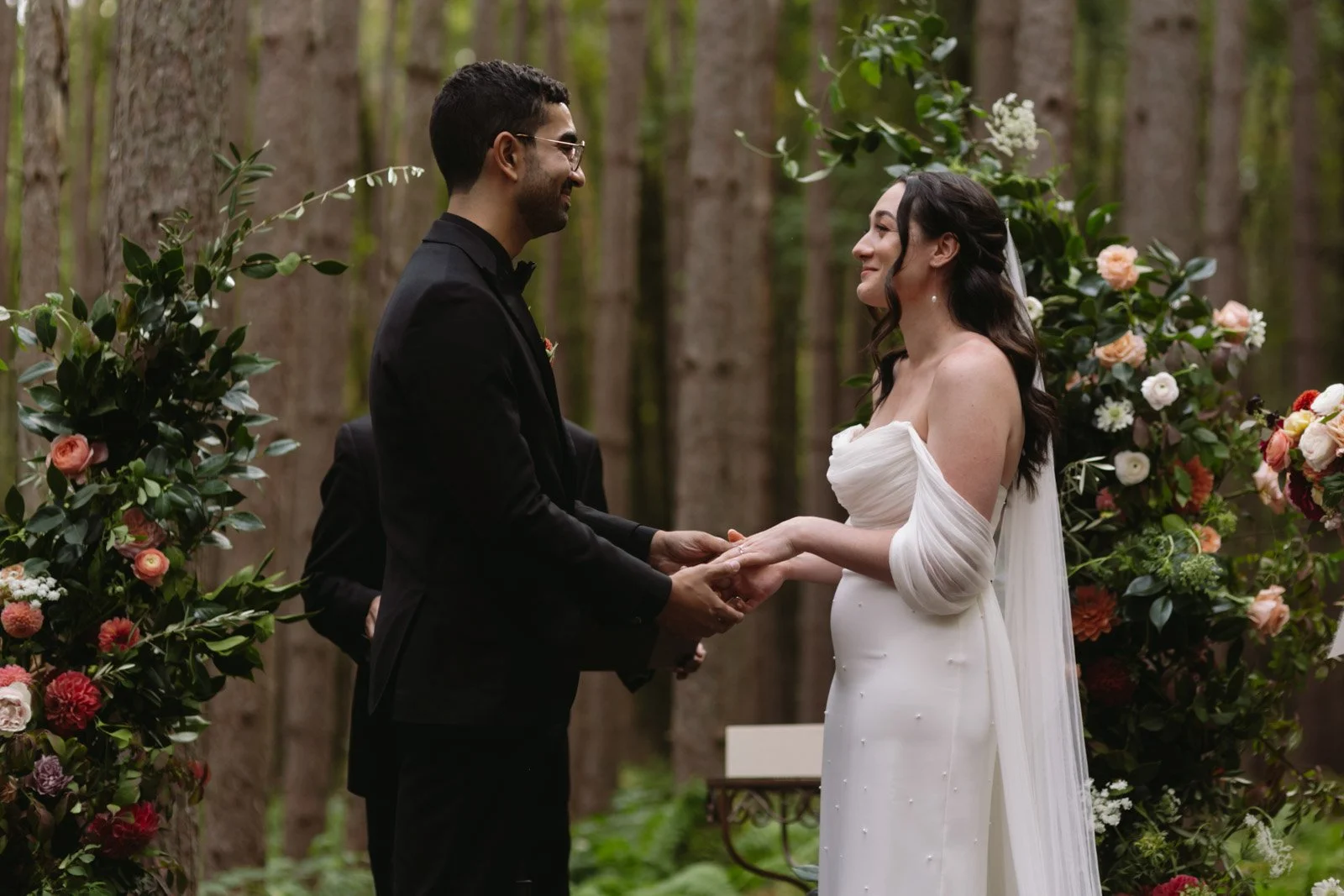 A bride and groom holding hands during a wedding ceremony in a forest, with floral decorations around them.