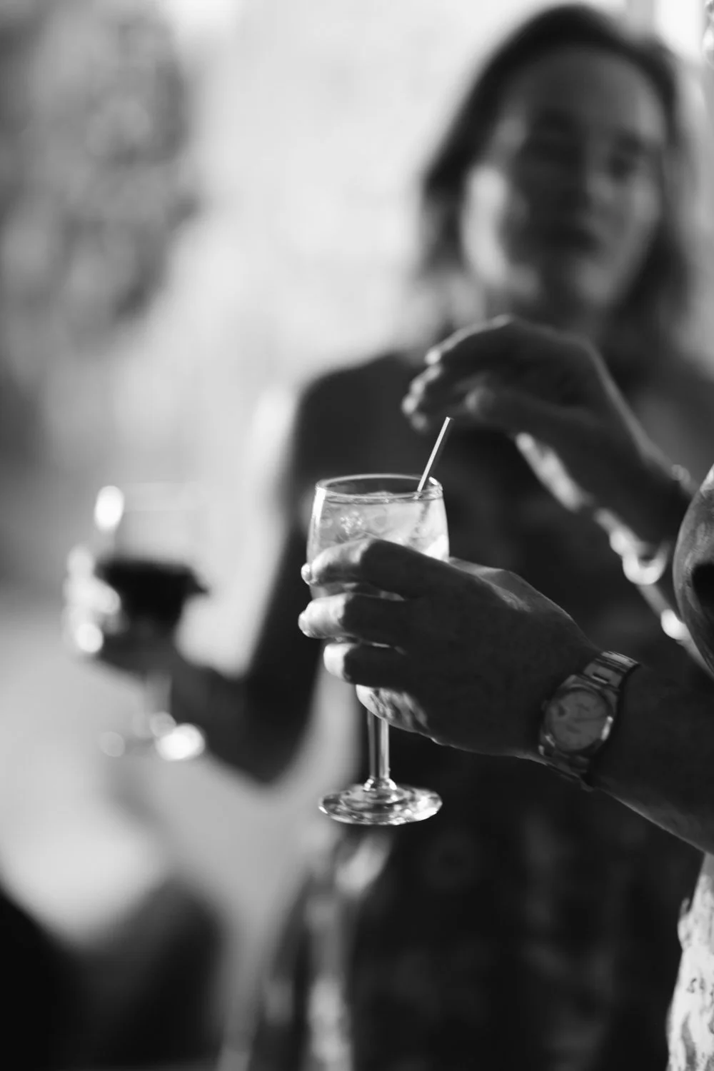 A person holding a glass of beverage with a straw, with another person in the background also holding a glass, in a social setting.