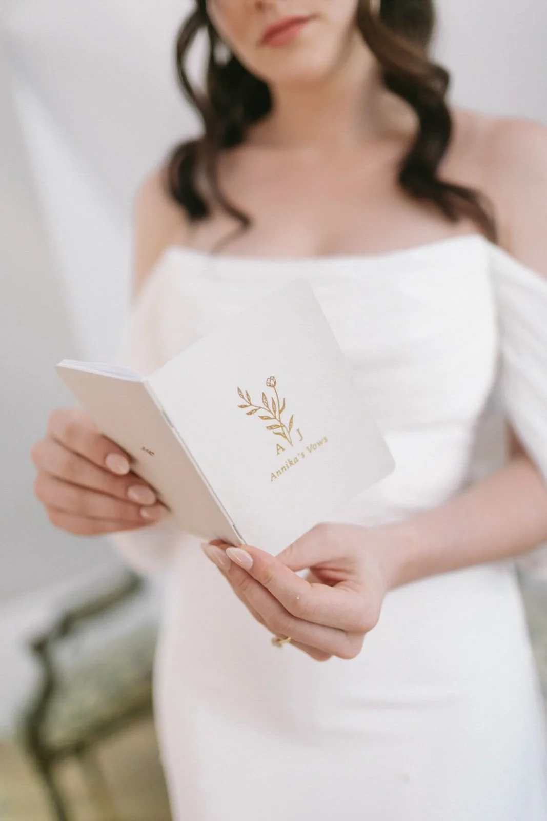 A woman in a white dress holding an invitation card with a gold logo and text reading 'Annika's Vows' and initials 'A J' with a floral design.