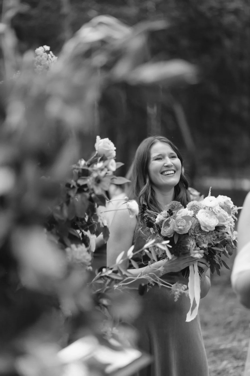 A woman smiling and holding a bouquet of flowers outdoors, surrounded by more flowers and foliage.