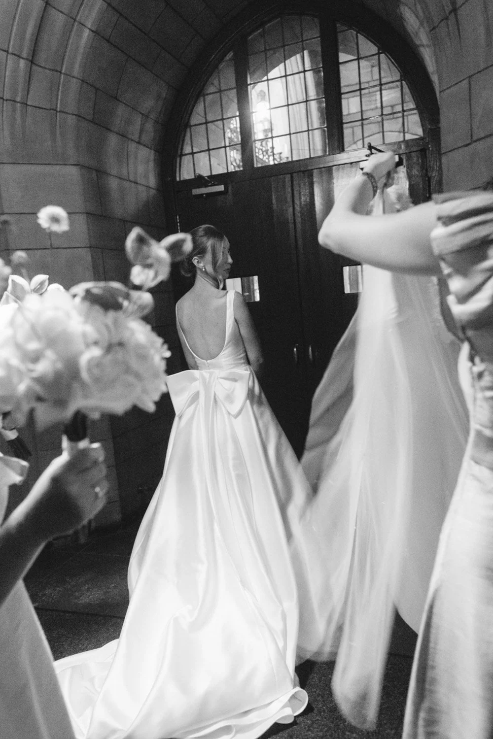 A woman in a wedding dress with a large bow back stands by a door in a dimly lit, arched wooden room, surrounded by other women in dresses, some holding bouquets.