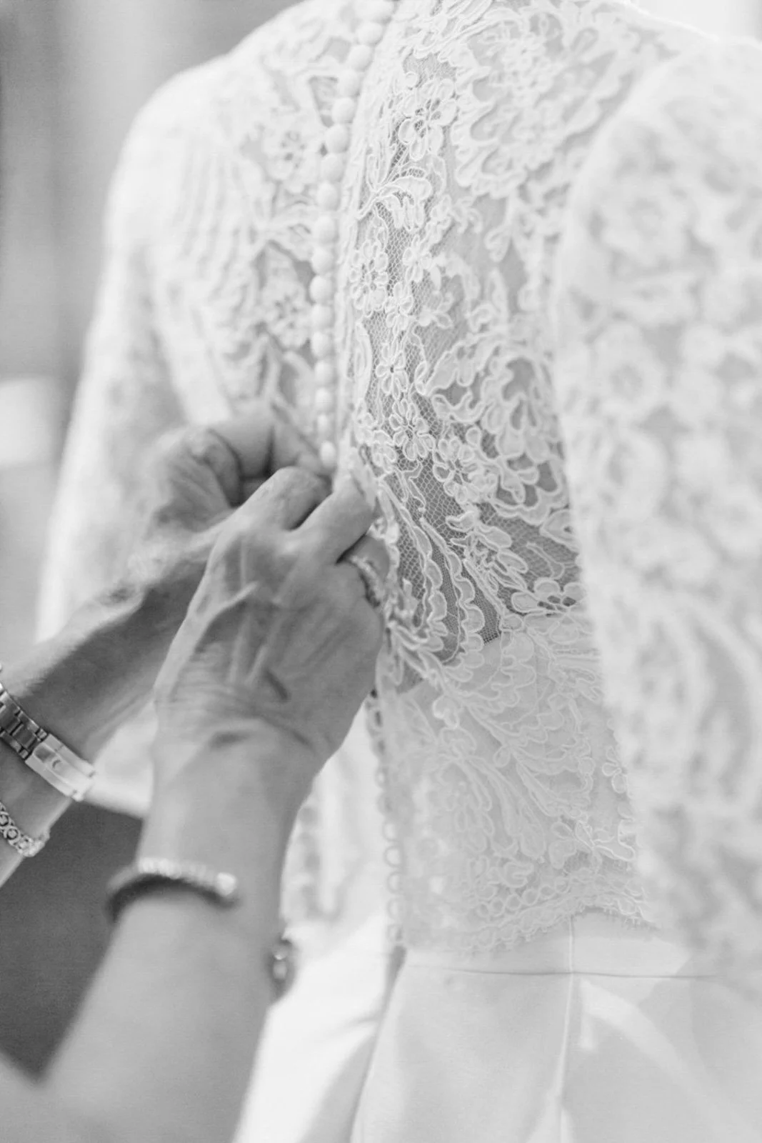 A woman adjusts a lace wedding dress with buttons on an intricately embroidered back.
