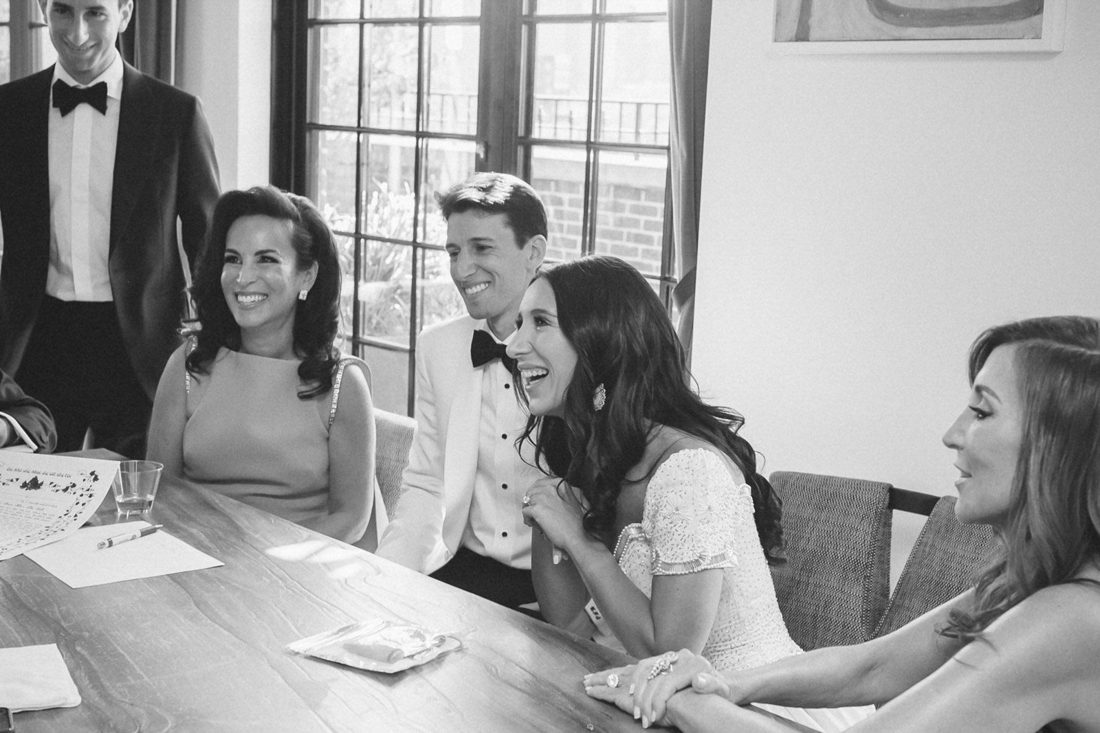 People sitting at a table, laughing and smiling during a social gathering or celebration. One woman in a white dress appears to be the bride, and others are dressed formally.