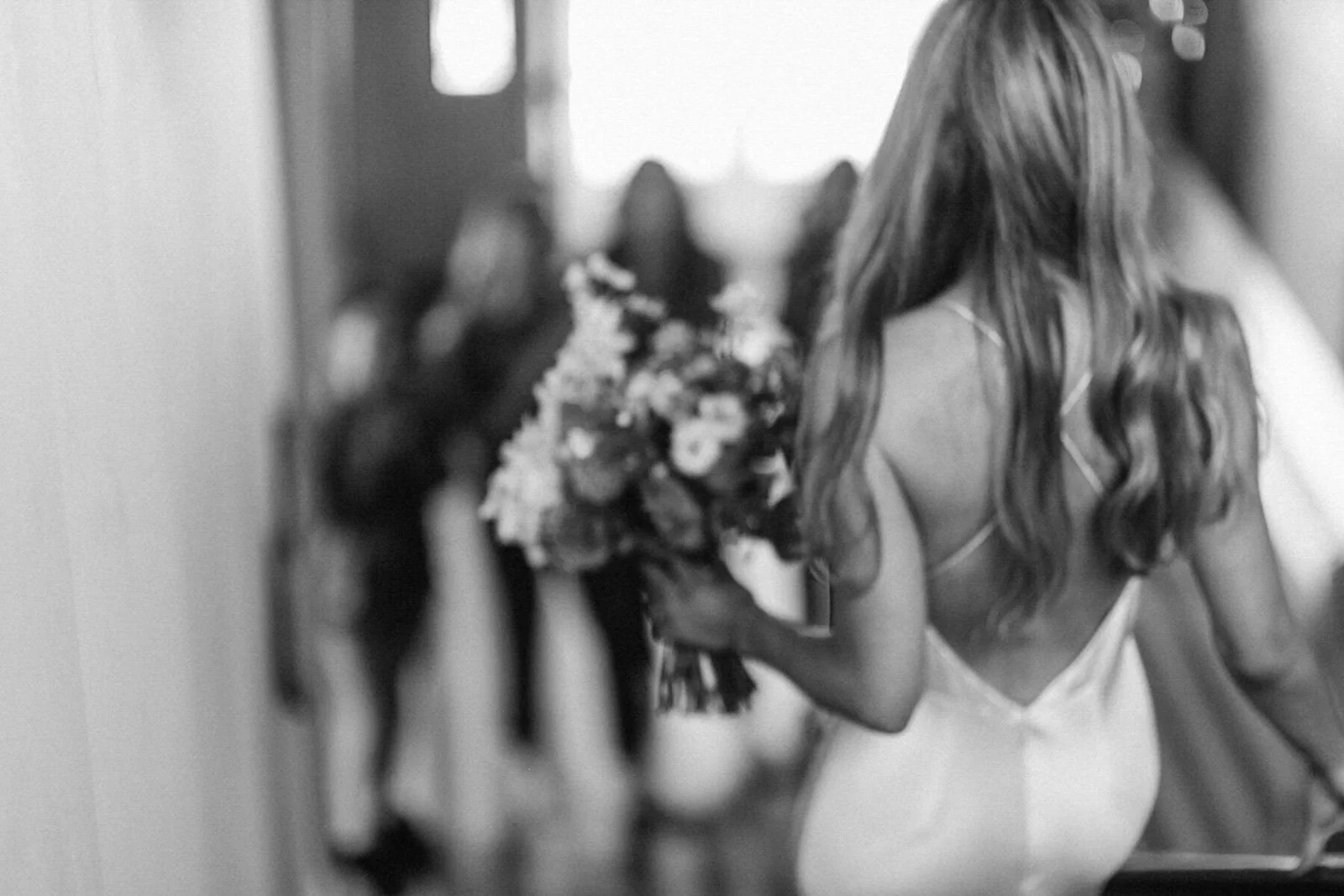 A woman in a white dress holding a bouquet of flowers, seen from the back, with three women in the background in a blurred setting.