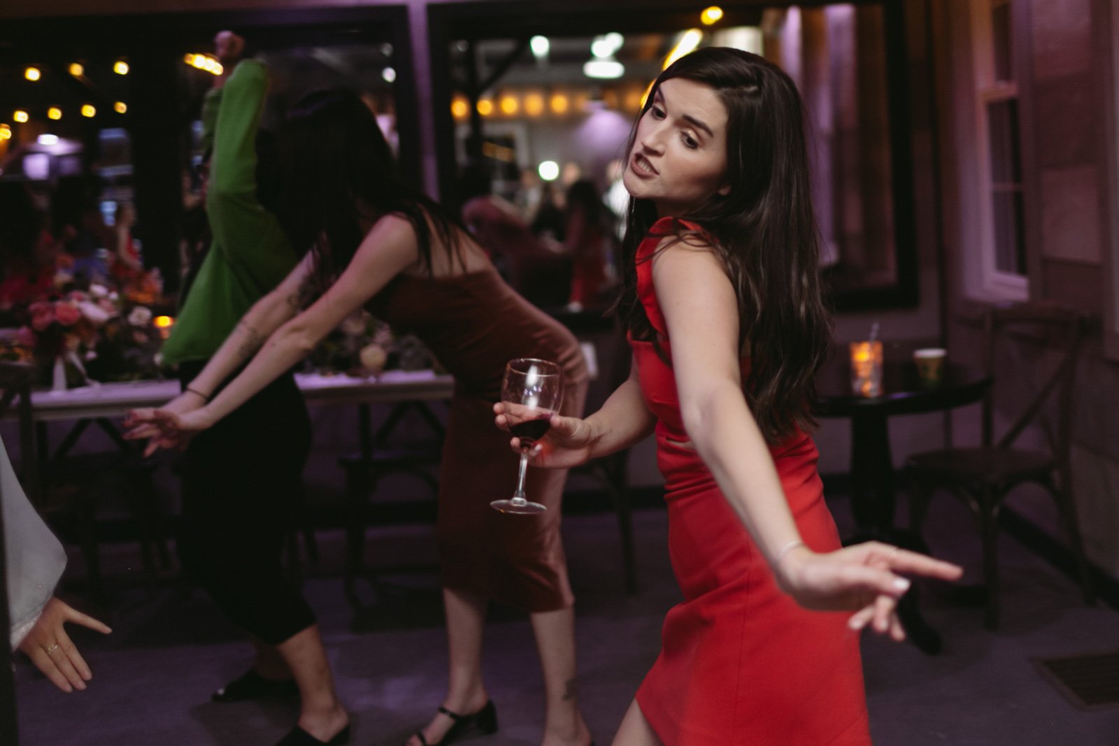A woman in a red dress dancing at a party holding a glass of red wine, with people dancing and socializing in the background.