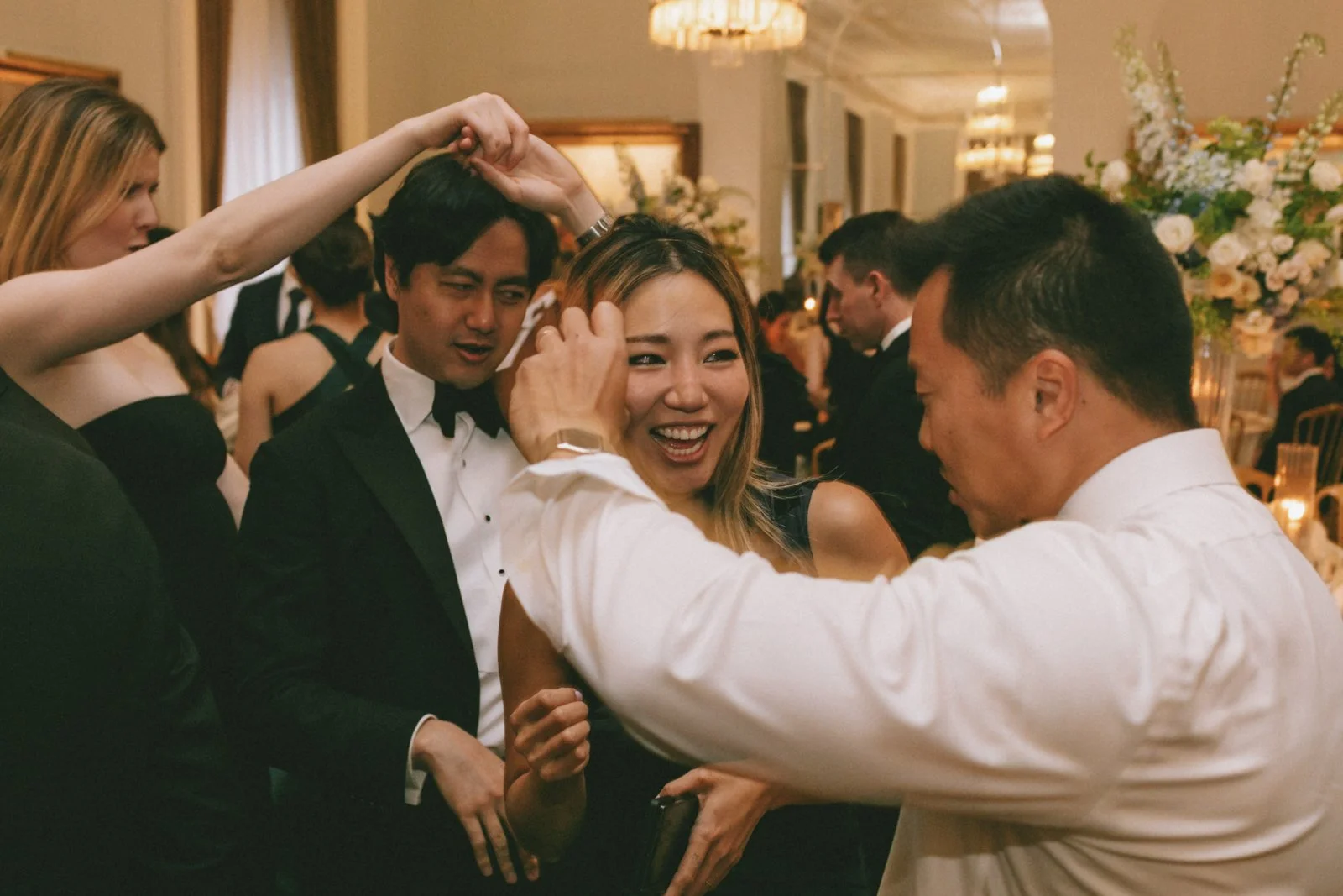 People dancing and socializing at a formal event, with a woman in a black dress smiling and a man in a tuxedo dancing with her.