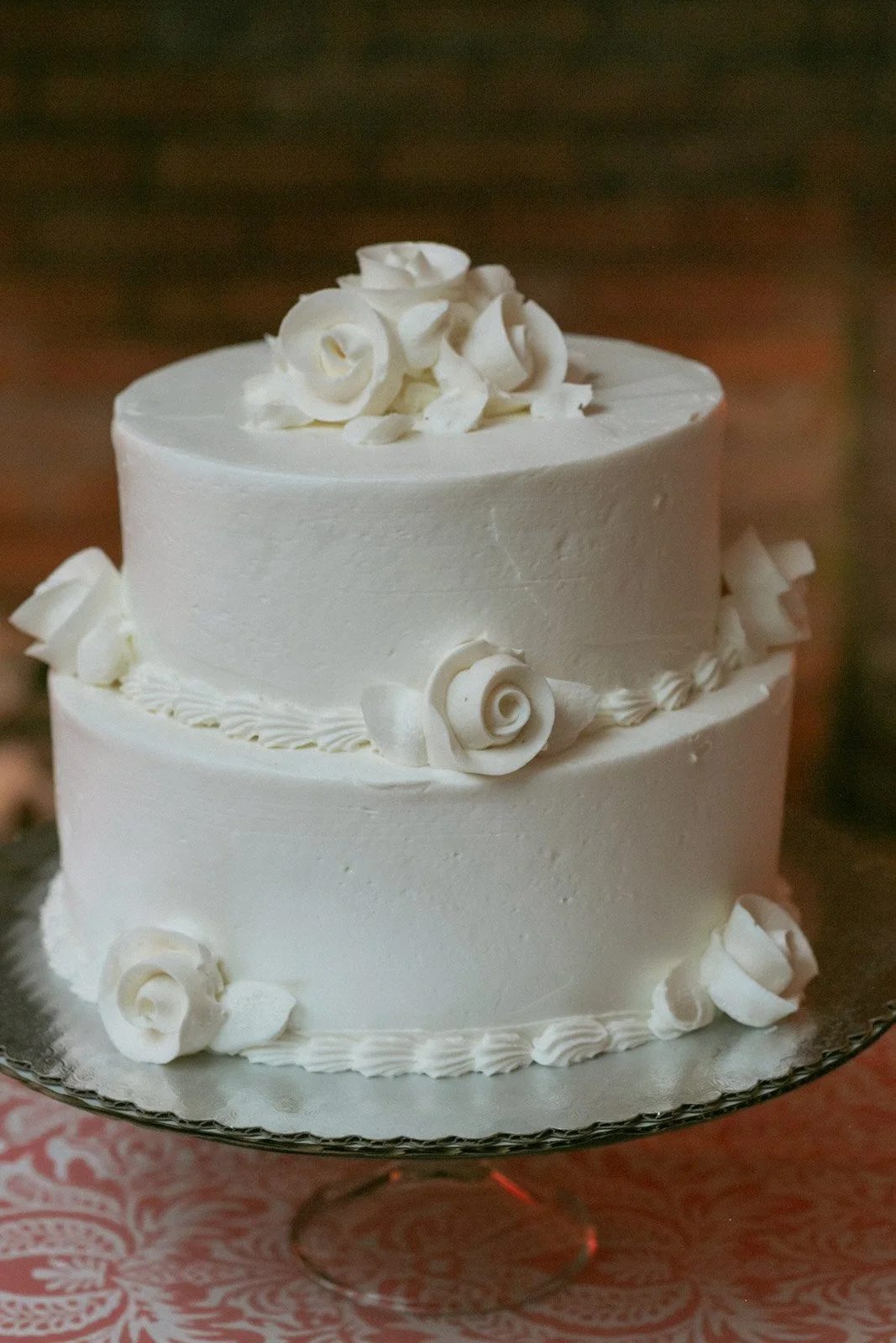 A two-tiered white wedding cake decorated with white roses and piped borders, placed on a glass cake stand.