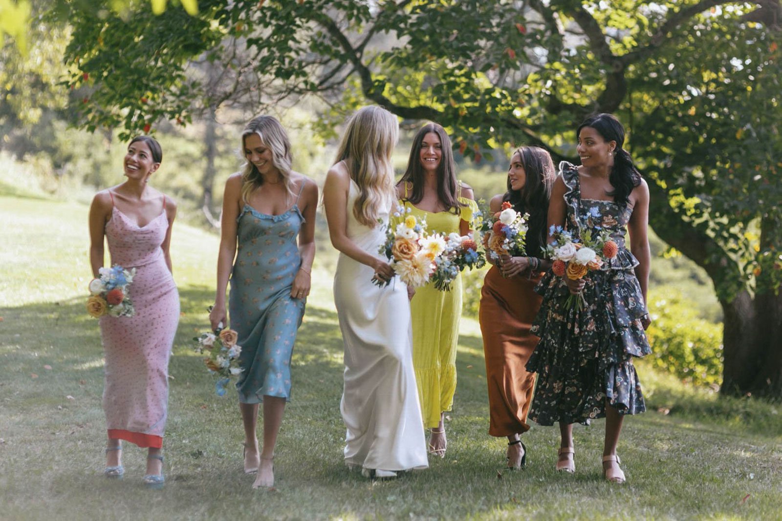 Bridesmaids walking outdoors on grass, holding bouquets of flowers, dressed in colorful dresses, smiling, with trees and sunlight in the background.