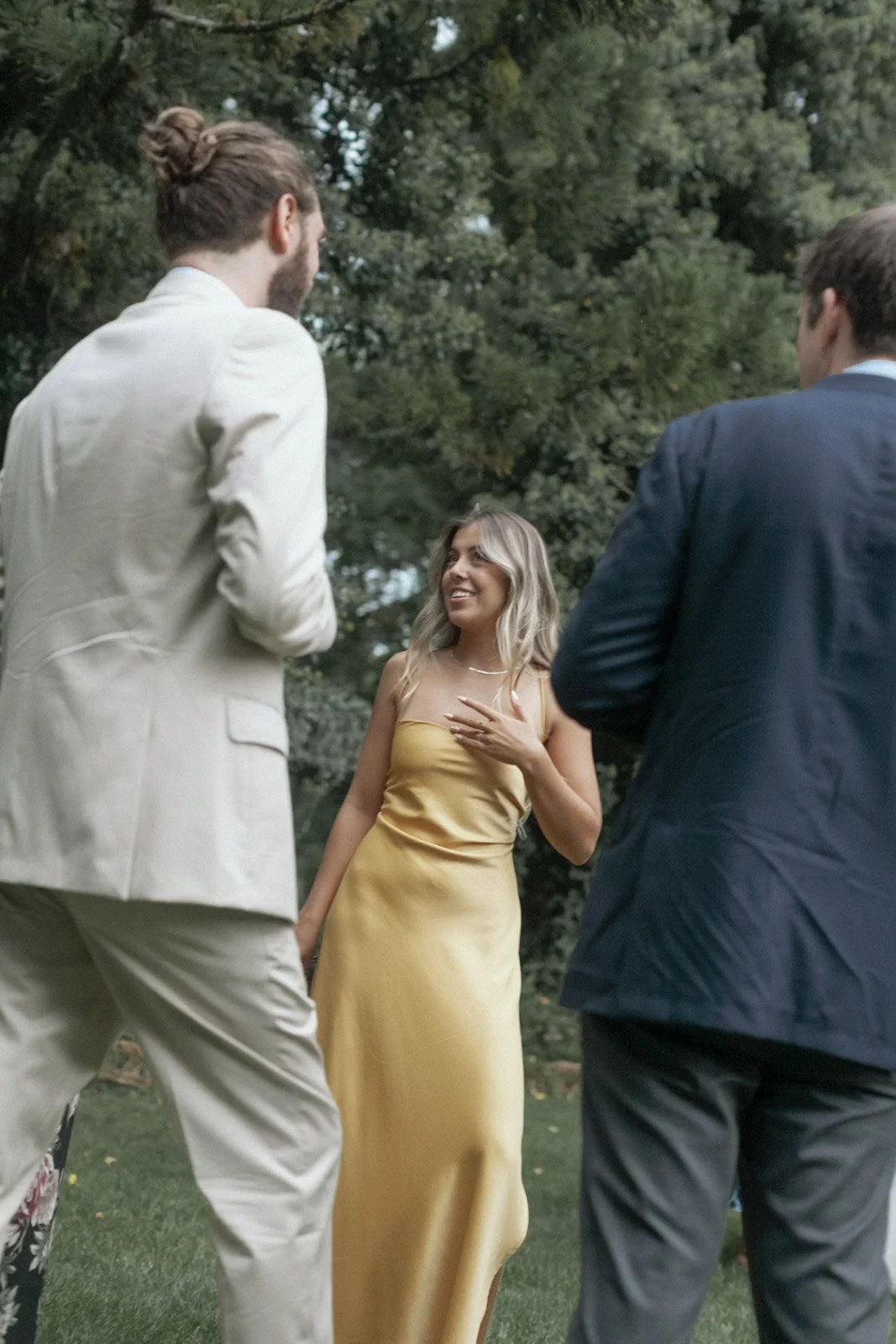 A woman in a yellow dress smiling and speaking to two men in formal suits outdoors with trees in the background.