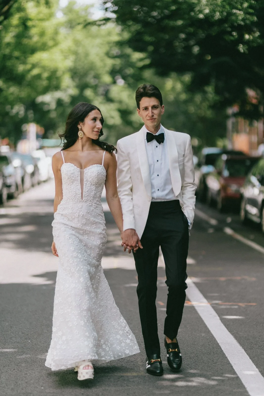 A couple in wedding attire walking hand in hand down a street lined with parked cars and green trees.
