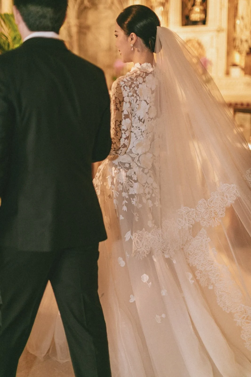 A bride in a lace wedding dress with a long veil, standing beside a groom in a black suit, during a wedding celebration.