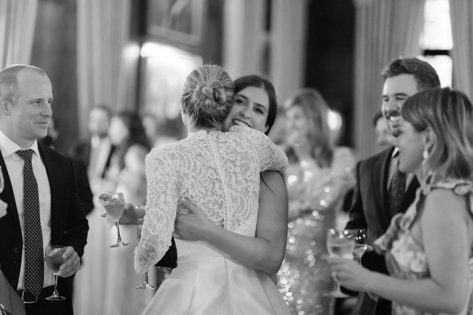 A woman in a wedding dress hugging another woman at a wedding reception, with several guests holding drinks and socializing in the background.
