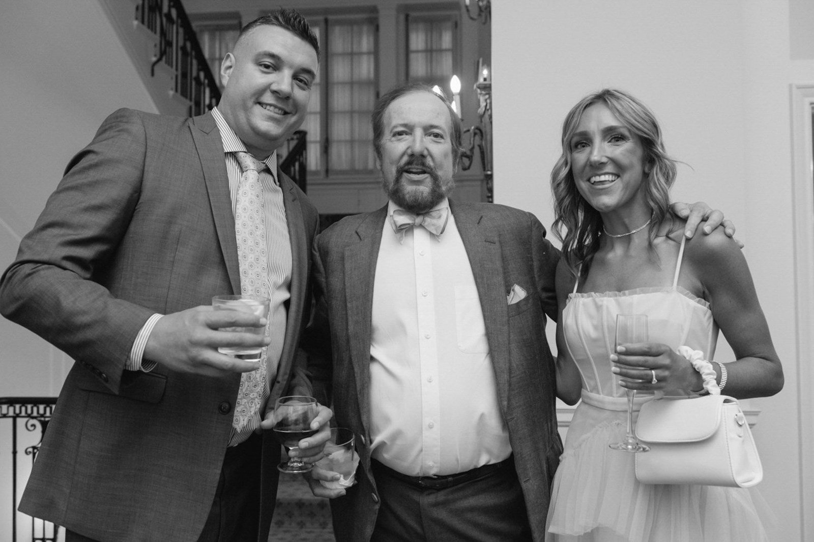 Black and white photo of three people at a social event, smiling and holding drinks in a well-decorated indoor setting.