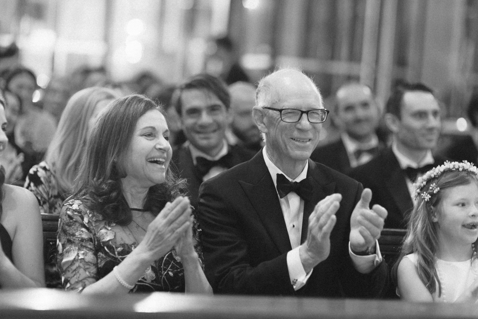 People at a gathering, clapping and smiling during an event, with a man in a tuxedo with glasses in focus.