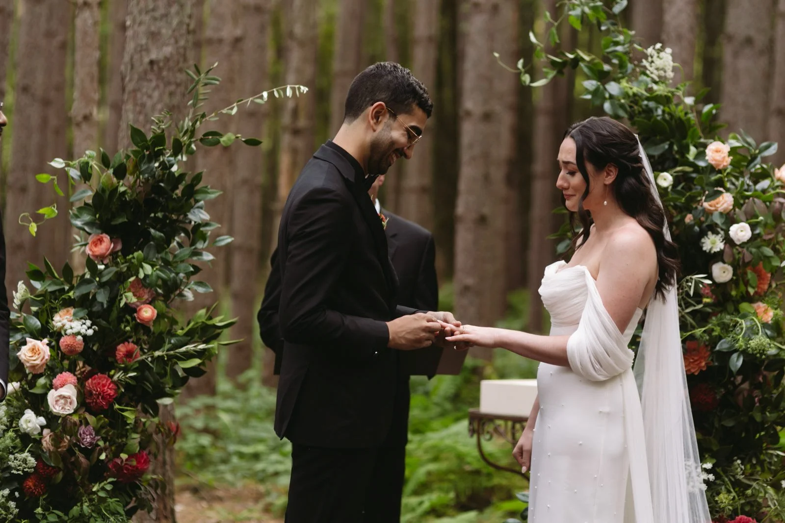 A couple getting married outdoors in a forest, exchanging rings, with floral arrangements on either side.