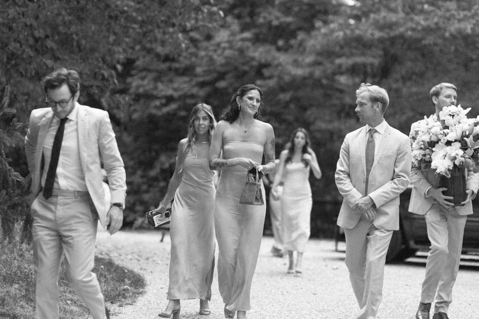 A group of six people dressed in formal attire walking outdoors on a gravel path, with trees in the background, including three women in dresses and three men in suits, one holding a large bouquet of flowers.