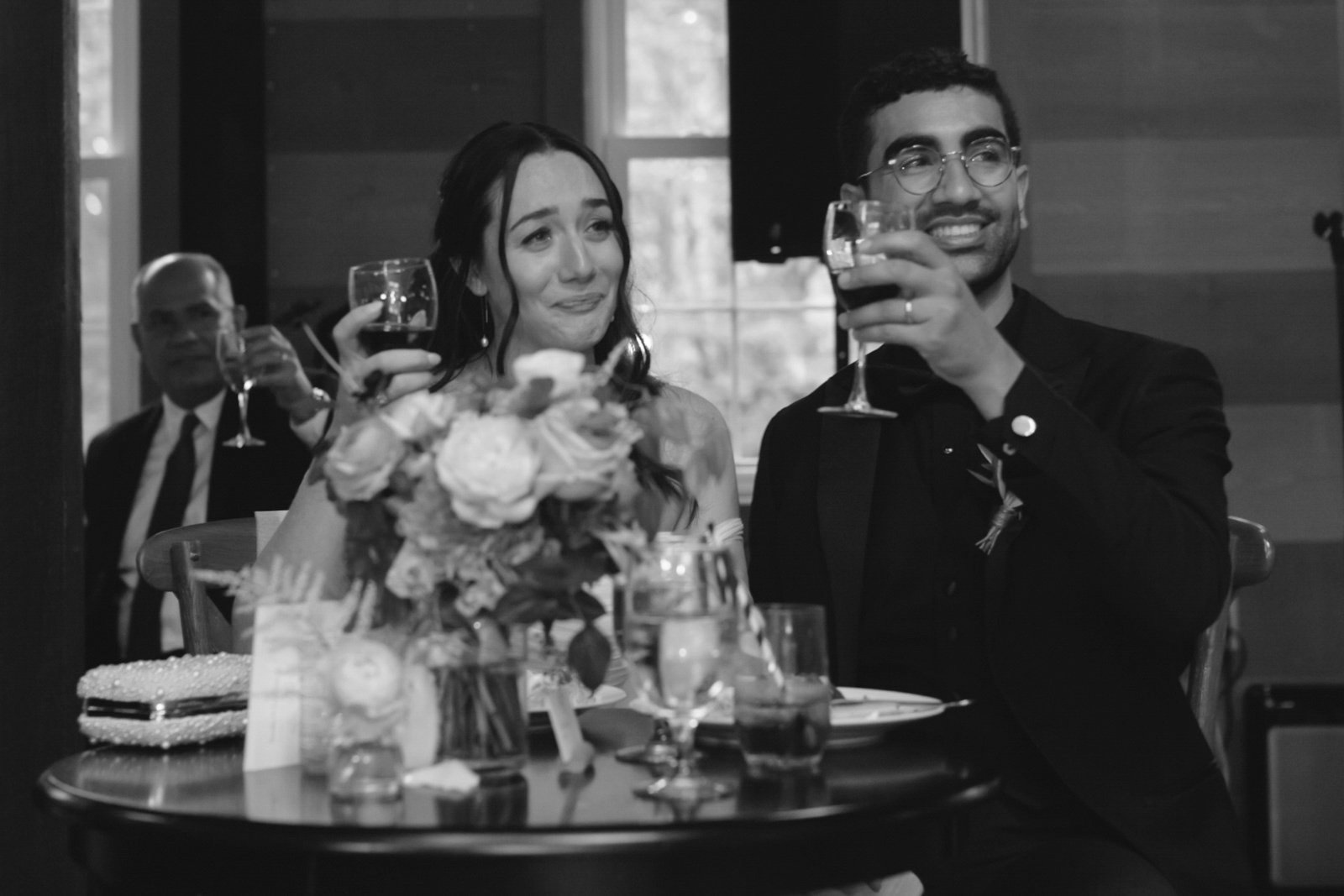 A black and white photo of a wedding reception, with a bride and groom holding glasses of wine and smiling, sitting at a decorated table with a bouquet of flowers, while a man in the background raises his glass in toast.
