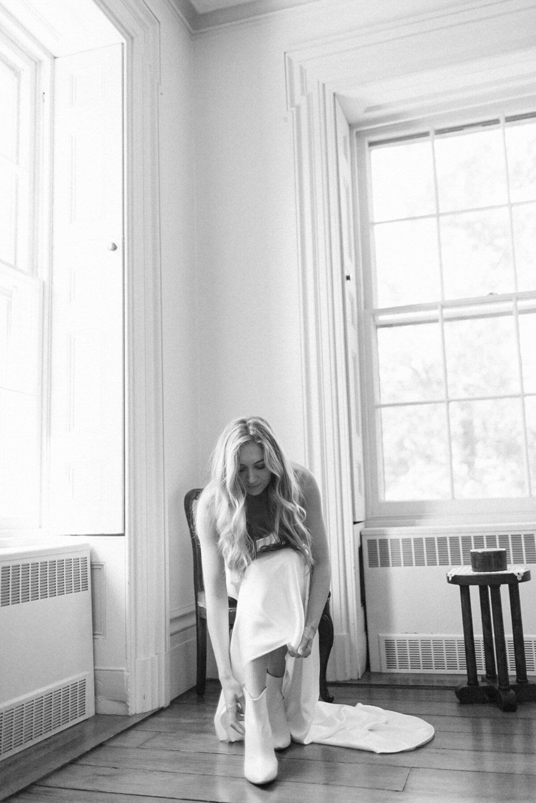 A bride with long wavy hair sitting on a chair in a room with large windows. She is putting on white boots while wearing a white dress, and there is a small table with a candle on it to her side.