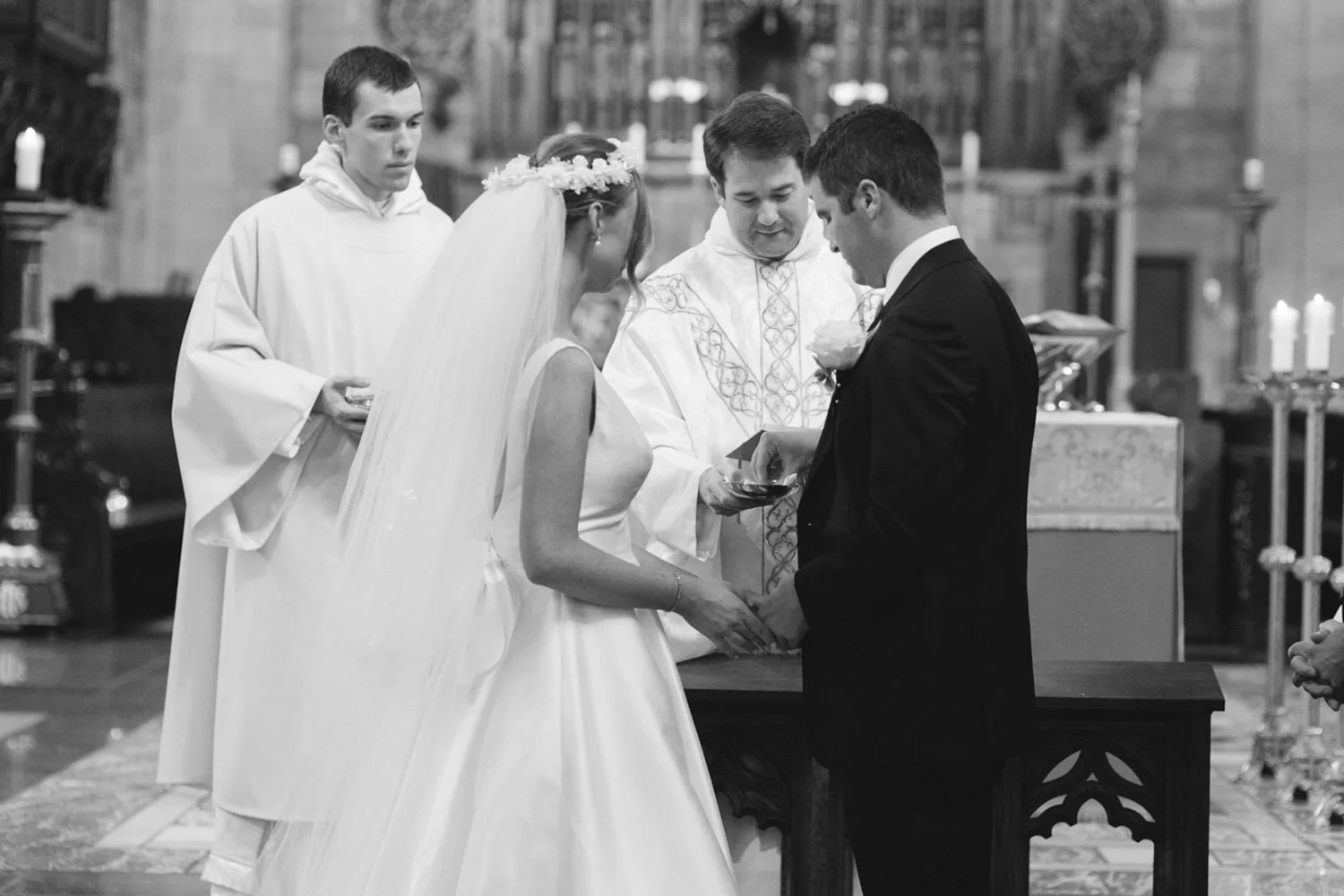 A black-and-white photo of a wedding ceremony inside a church, with the bride and groom holding hands and looking down, surrounded by two officiants. The bride is wearing a traditional white wedding dress with a veil and floral headpiece, while the g