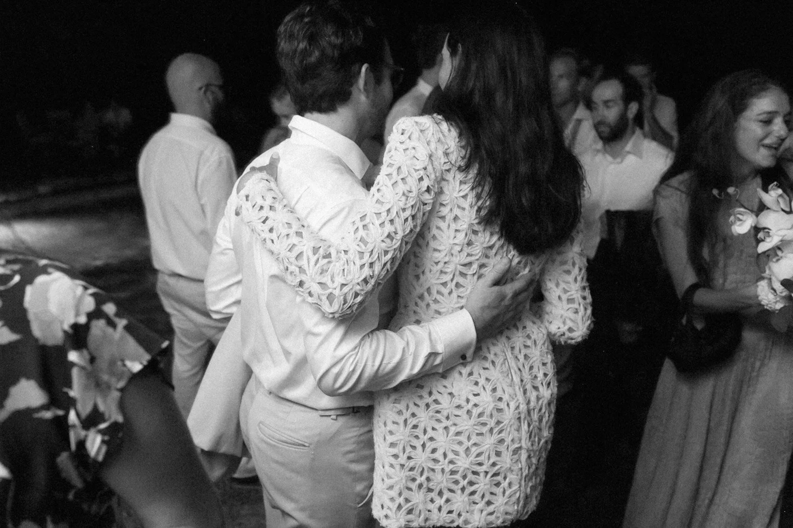 Black and white photo of a couple dancing closely at a social event, surrounded by other people, some holding flowers.