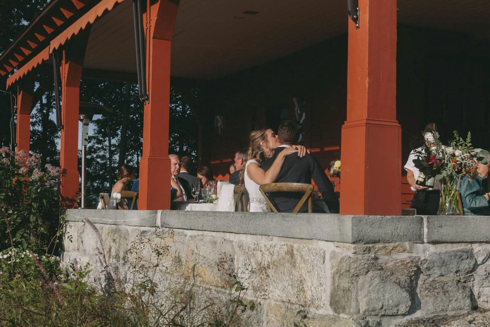 A couple sharing a kiss at a wedding reception outdoors under a red wooden pavilion, with several other guests seated at tables nearby.