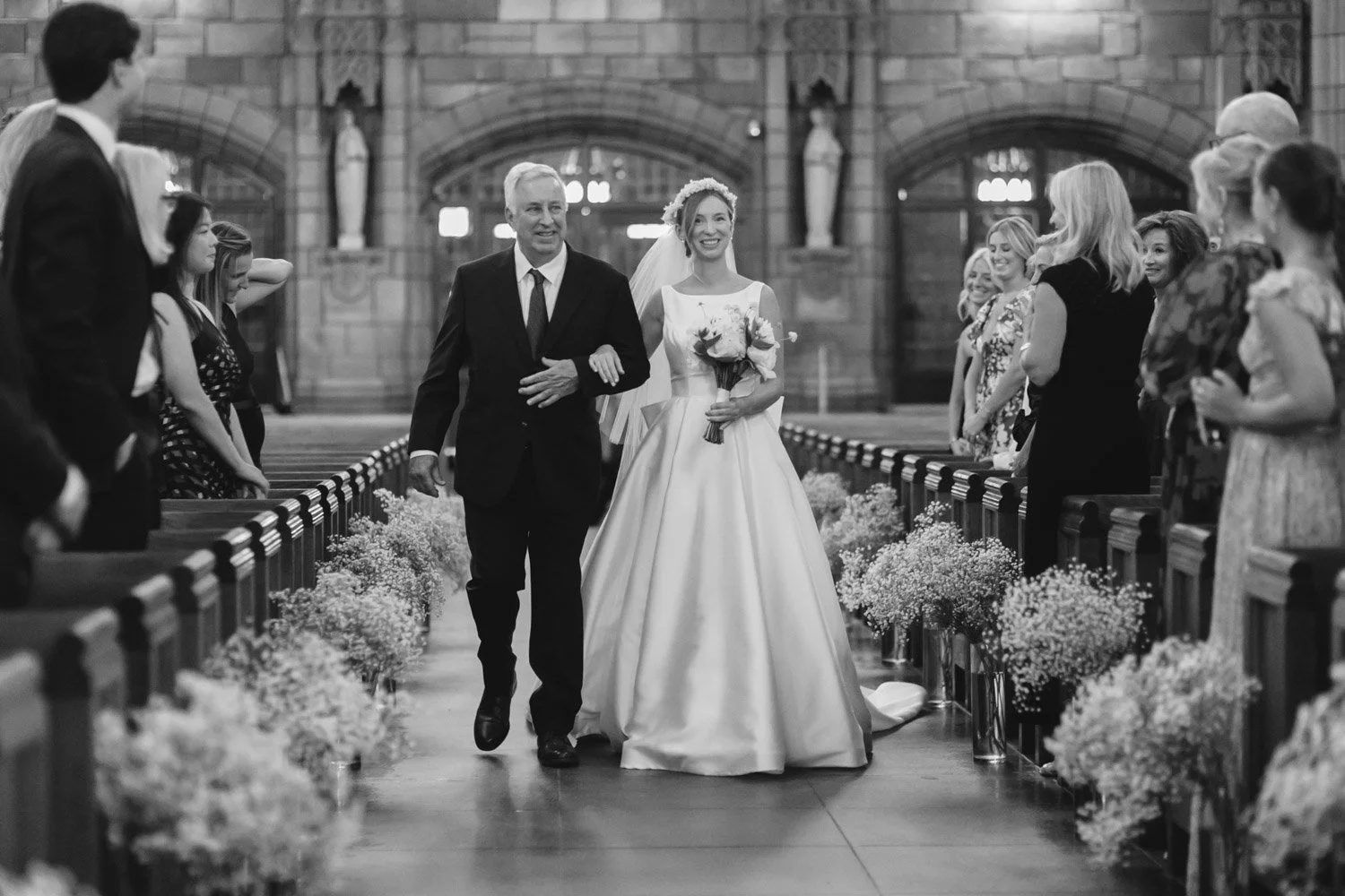 Bride walking down the aisle with her father in a church, smiling, holding a bouquet, surrounded by guests.