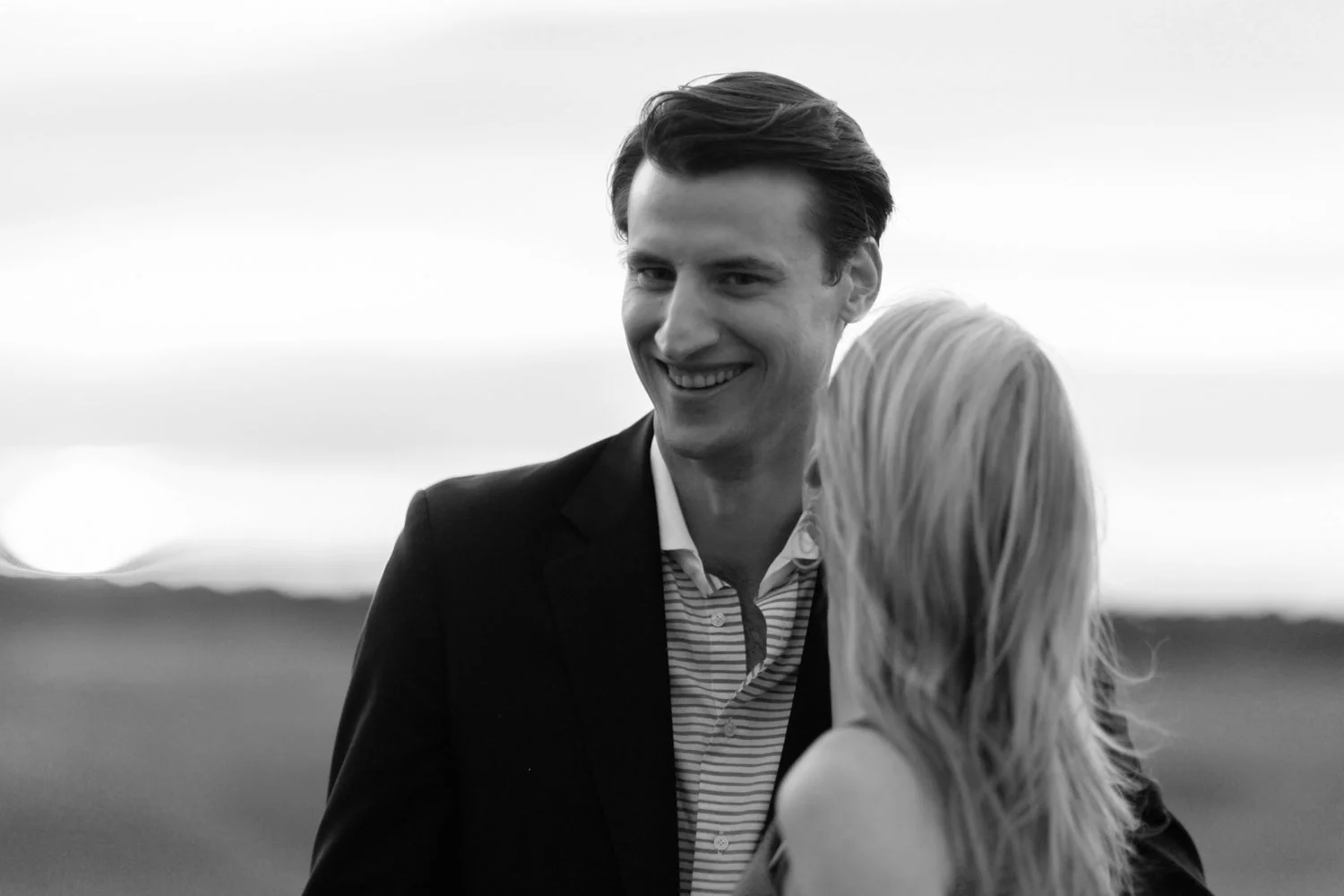 A black and white photograph of a smiling man in a suit talking to a woman with long blonde hair outdoors.