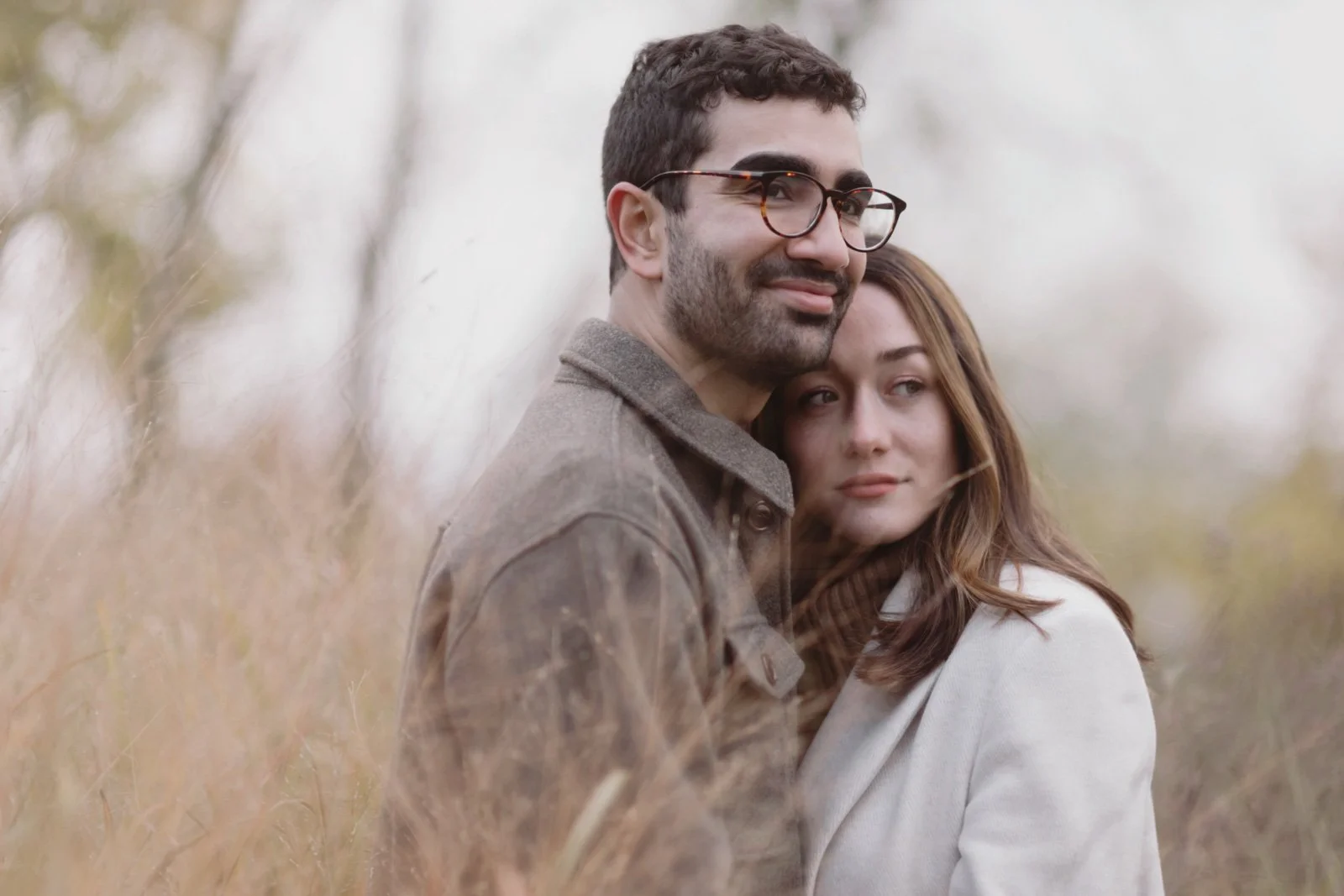 A couple standing close together outdoors in a field of tall, beige grass, smiling softly and looking at the camera, with a blurred background of trees and sky.