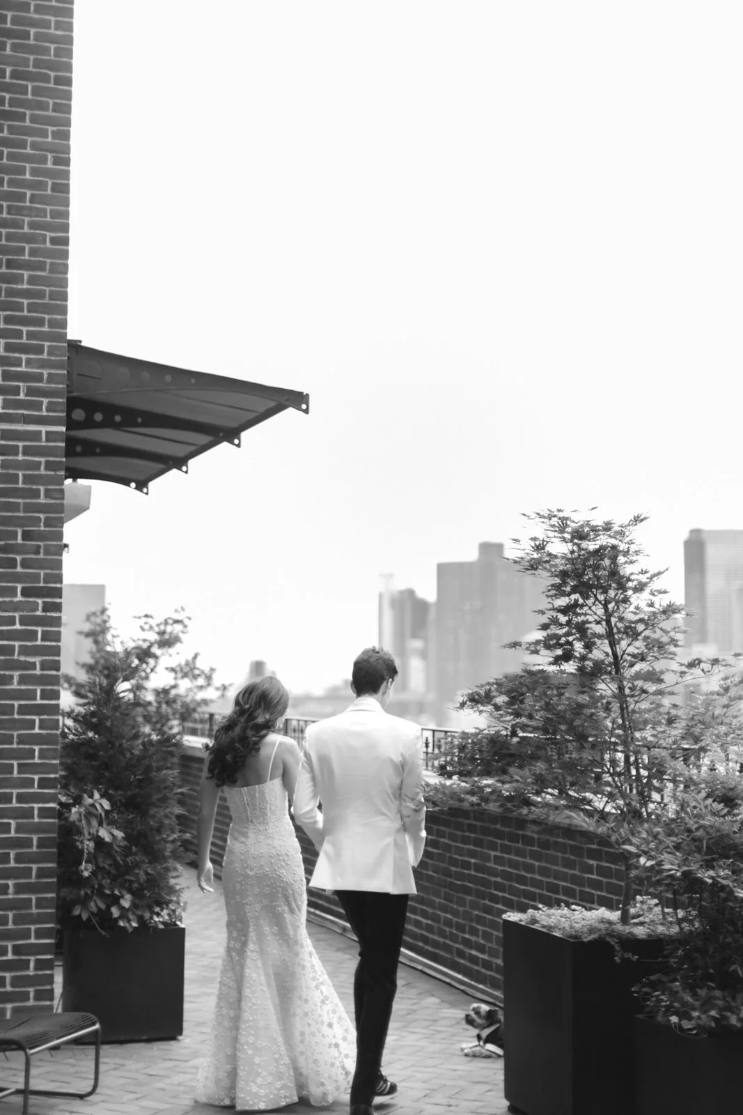 A couple, dressed in formal attire, holding hands while walking on a rooftop terrace with potted plants and city skyline in the background.