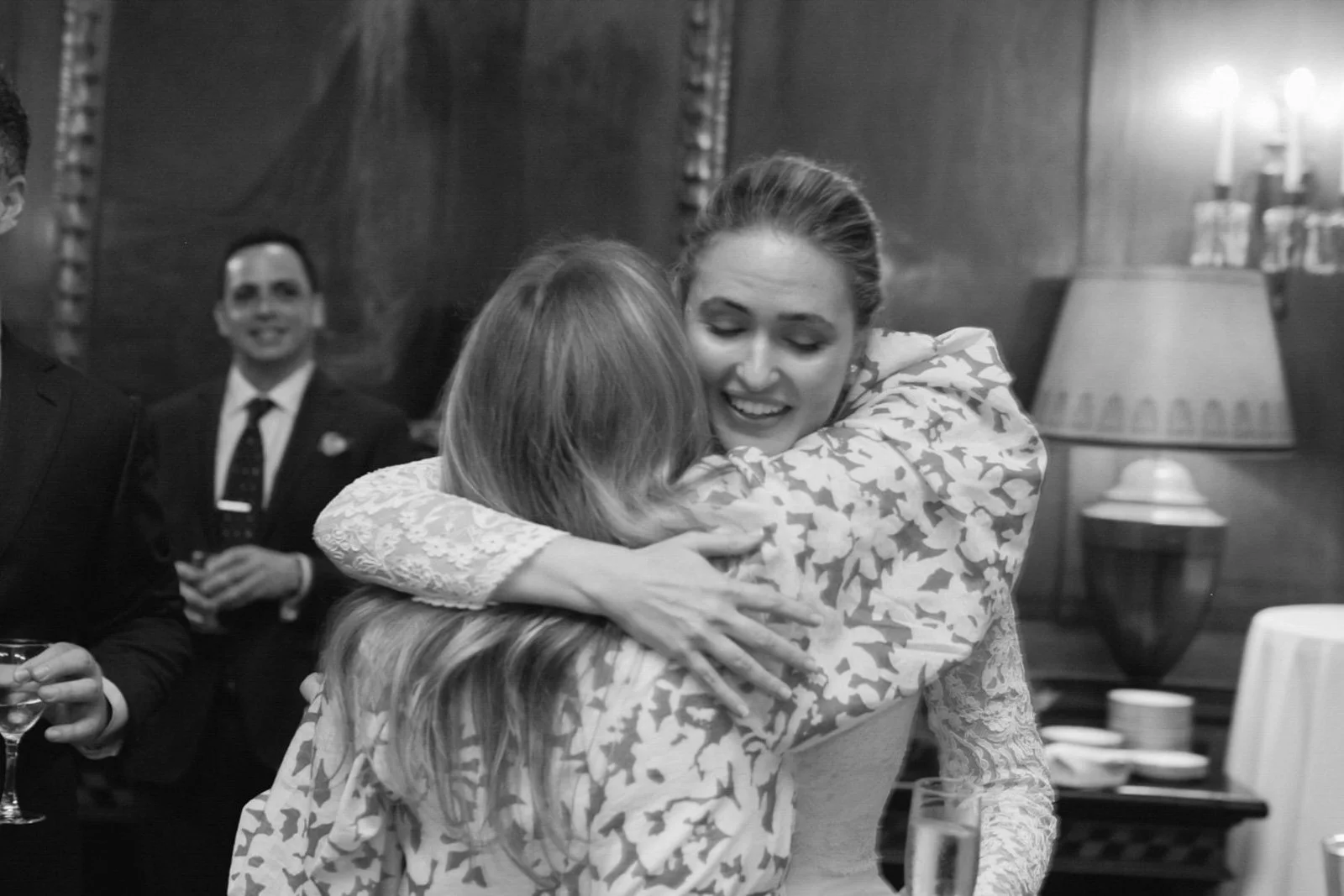 Two women hugging at a social gathering, with other people in the background smiling and holding drinks.