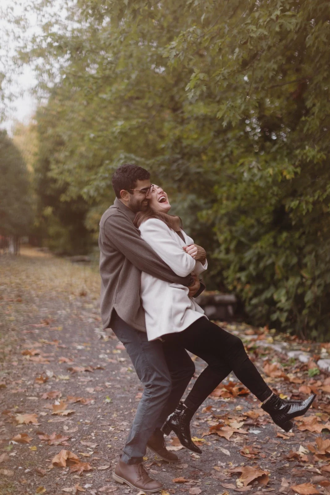 A man and woman laughing and hugging outdoors in autumn, walking on a leaf-covered path among trees.