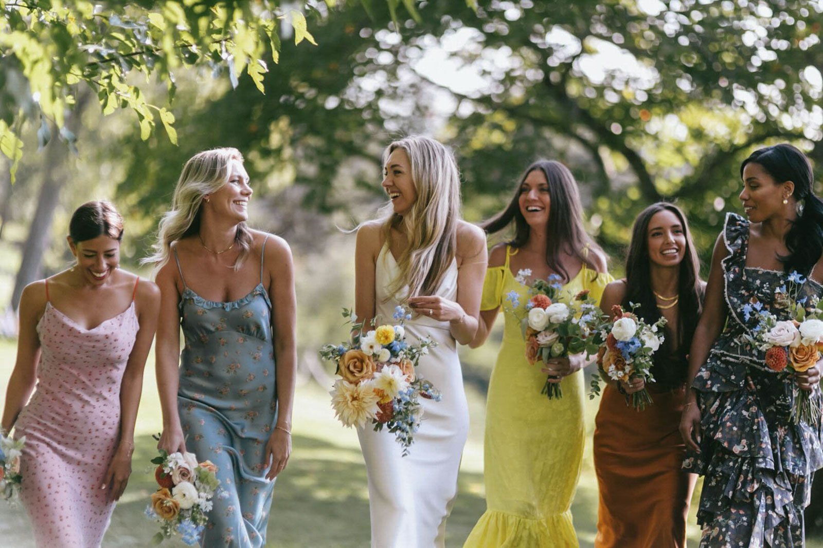 Bridesmaids dressed in colorful dresses, holding bouquets of flowers, walking outdoors and smiling.