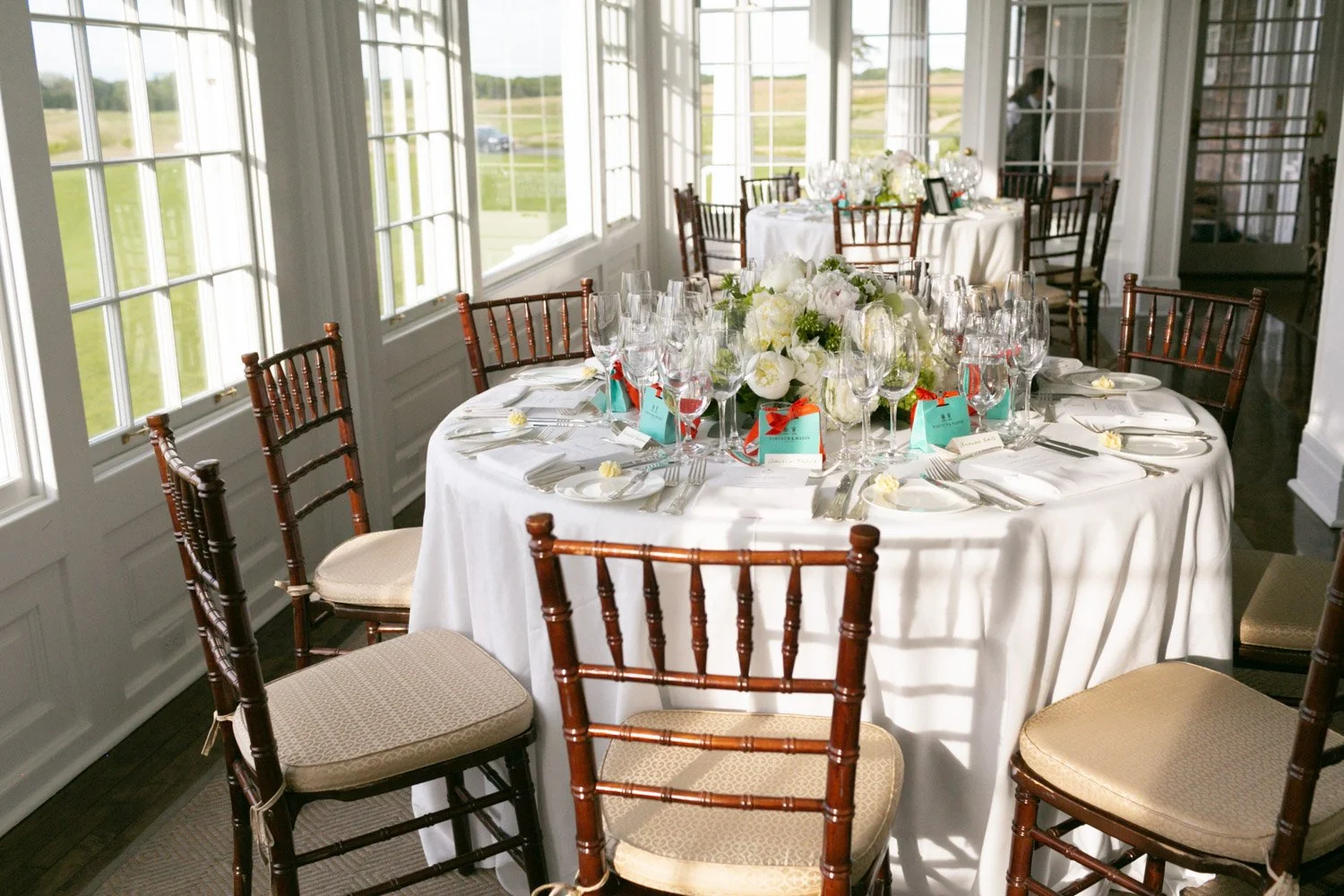 Round table set for a wedding reception with white tablecloth, floral centerpiece, and multiple wine glasses and cutlery, surrounded by wooden chairs in a sunlit room with large windows.