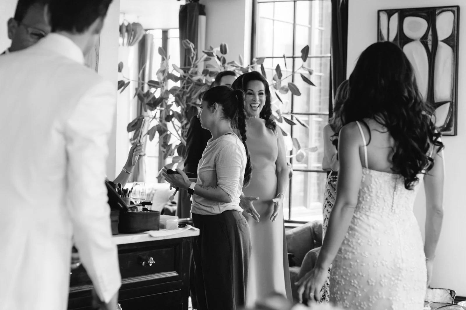A group of women and a man in a bright room, some women in casual clothes, one woman in a white dress, possibly preparing for a wedding. They are smiling and chatting, with makeup and beauty products visible on a table.