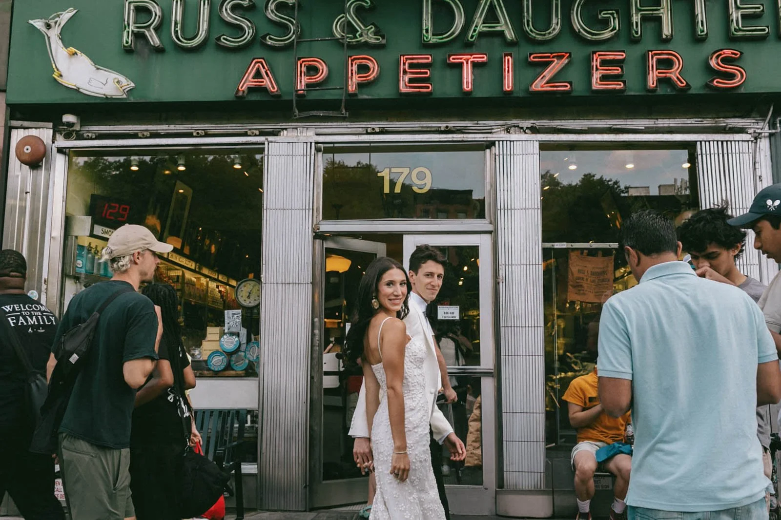 People standing outside a restaurant with a neon sign that reads 'Russ & Daughters Appetizers'. Two women in the center, one in a white dress, smiling at the camera. Several other people are gathered around, some talking or looking at their phones, w