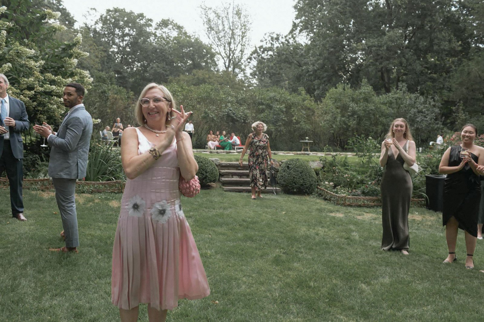 People gathered outdoors in a garden, dressed in formal attire, celebrating and clapping. A woman in a pink dress is smiling and clapping, while others around her are enjoying the event.