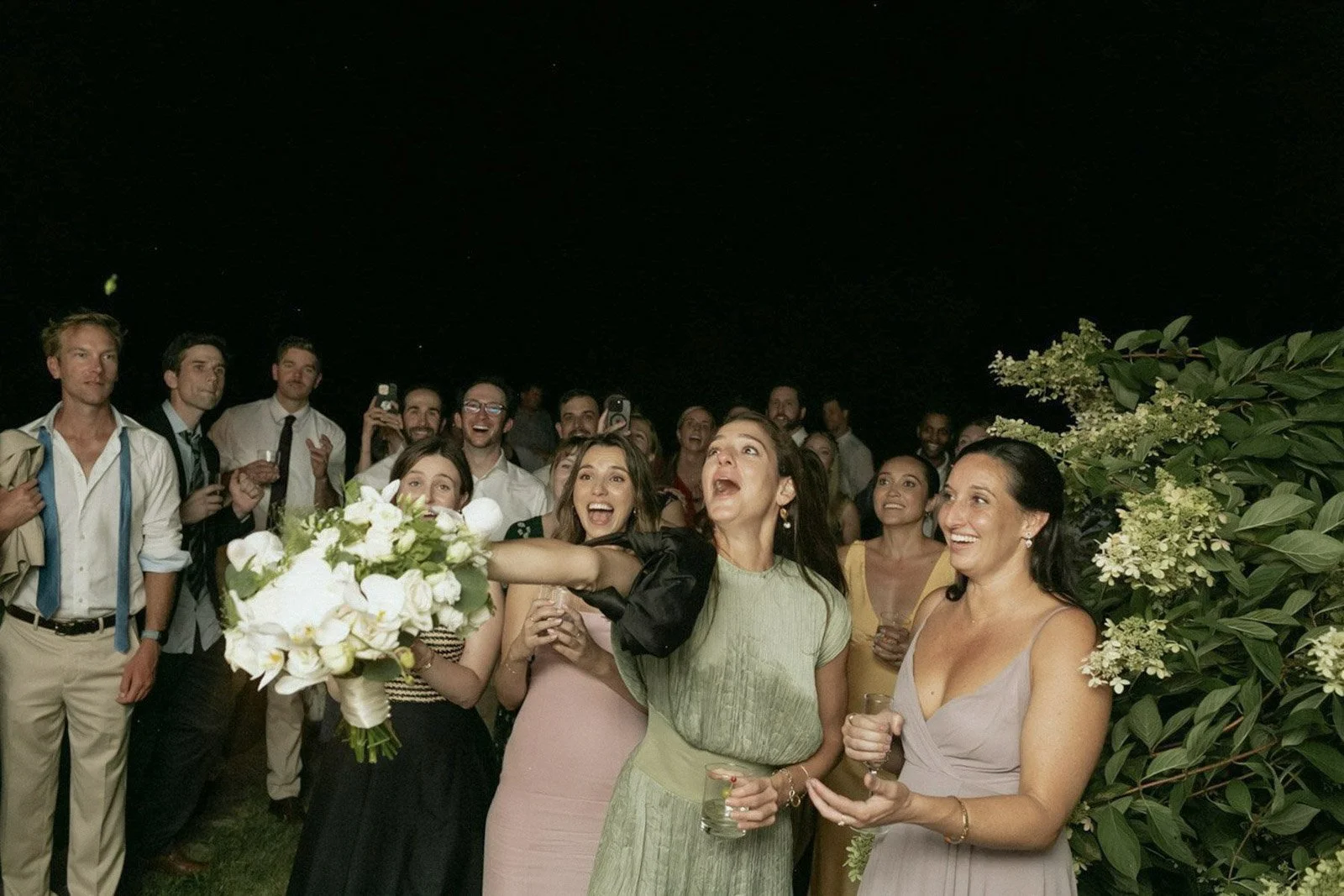 Group of people celebrating at a wedding reception outdoors at night, with women holding drinks and smiling while a woman throws a bouquet.