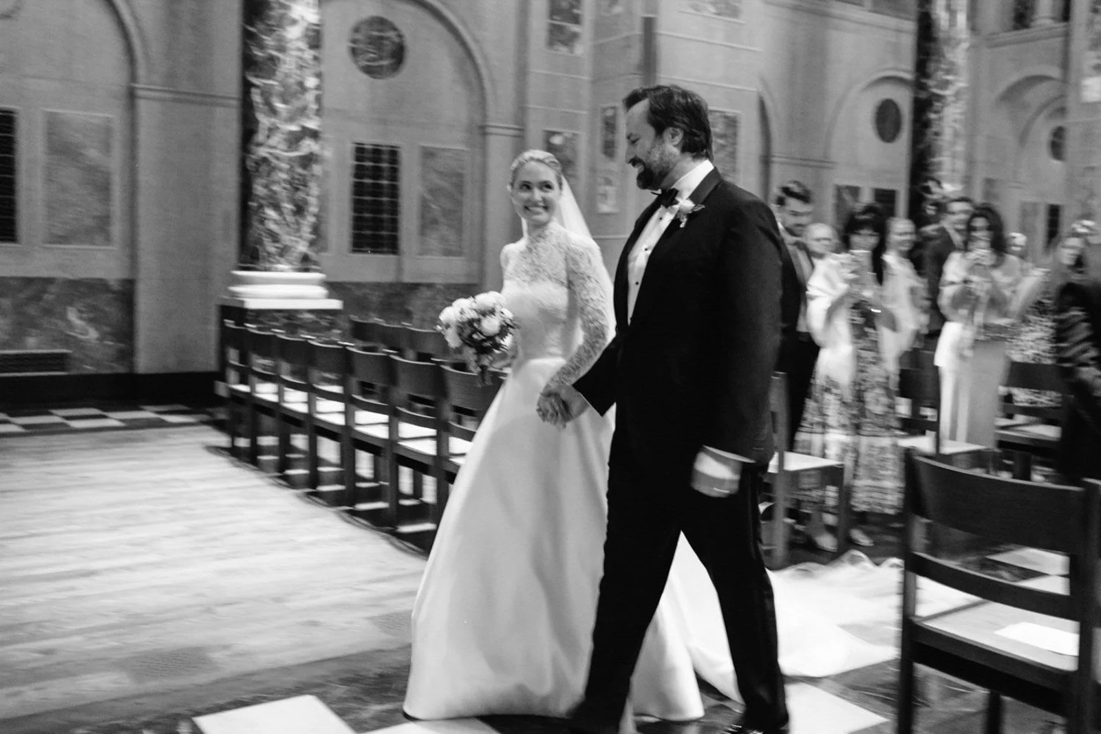A bride in a white dress holding a bouquet and a groom dressed in a black tuxedo walking down the aisle in a church, surrounded by seated guests.