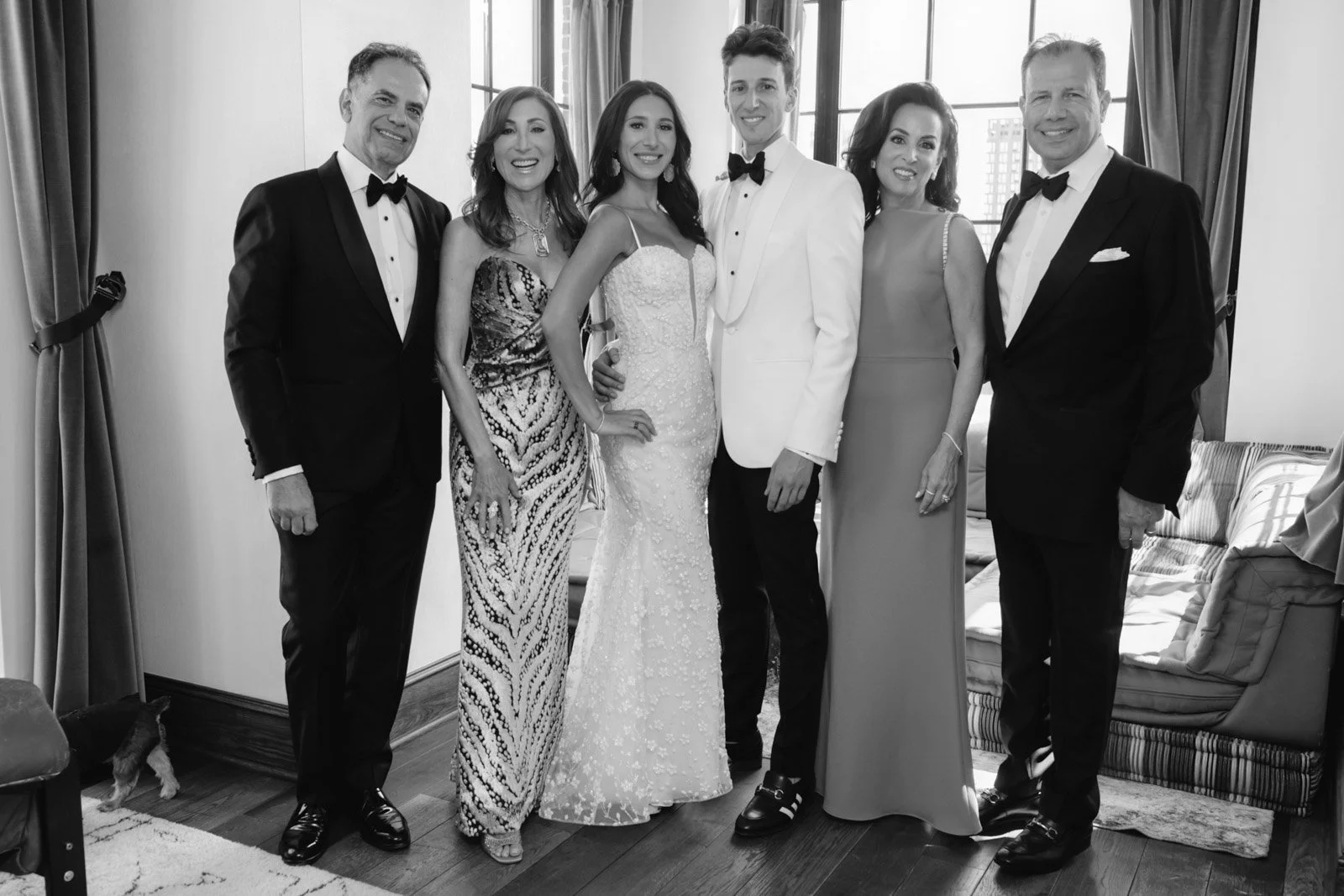 A wedding group photo of seven people in formal attire, including a bride and groom, standing indoors in front of large windows.