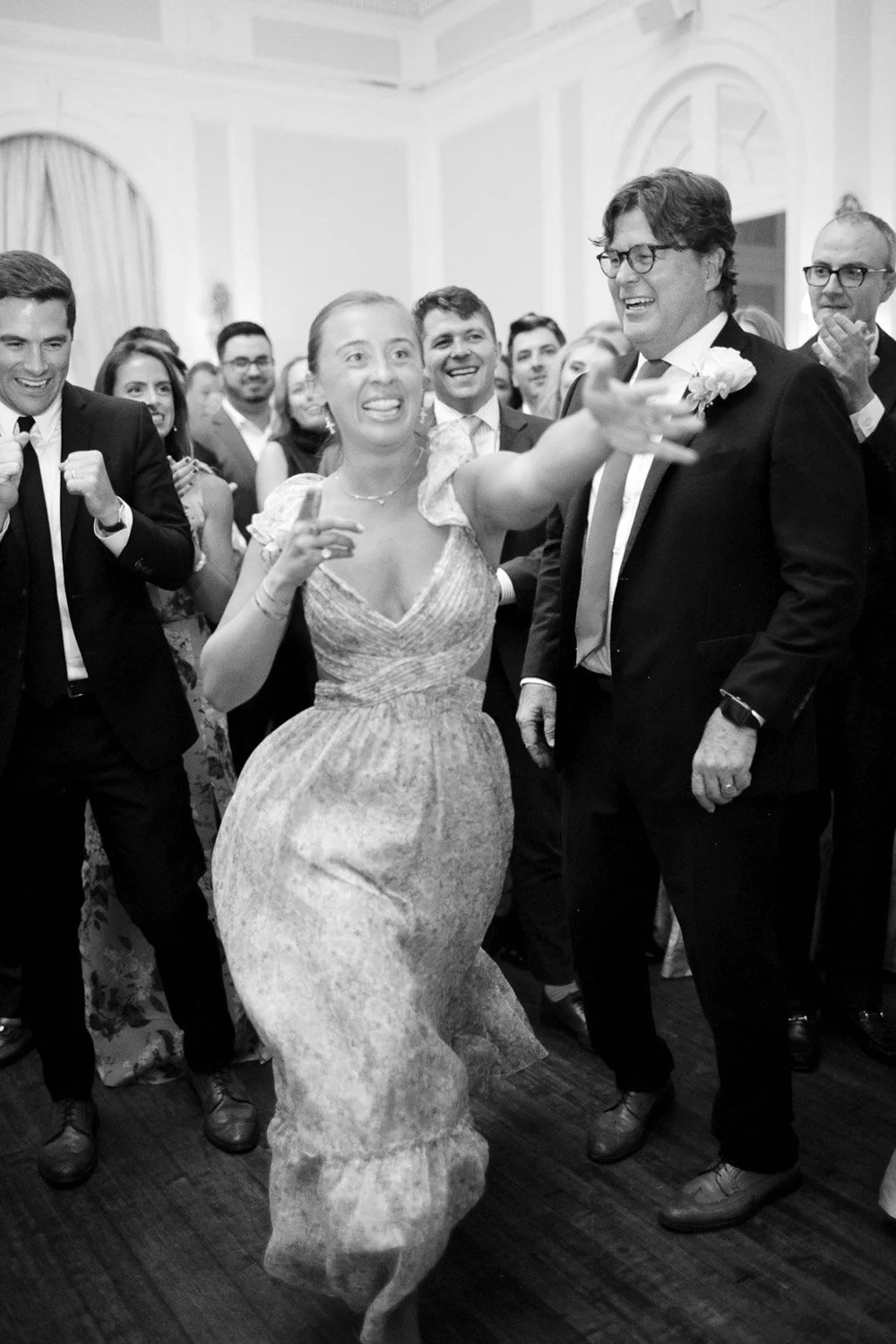 A woman in a dress is dancing with a man in a suit at a wedding reception, surrounded by smiling guests.